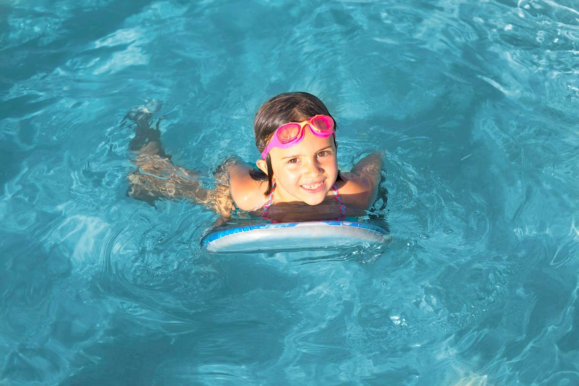 A young girl swimming in a pool with pink goggles, smiling at the camera, riding on a small float.