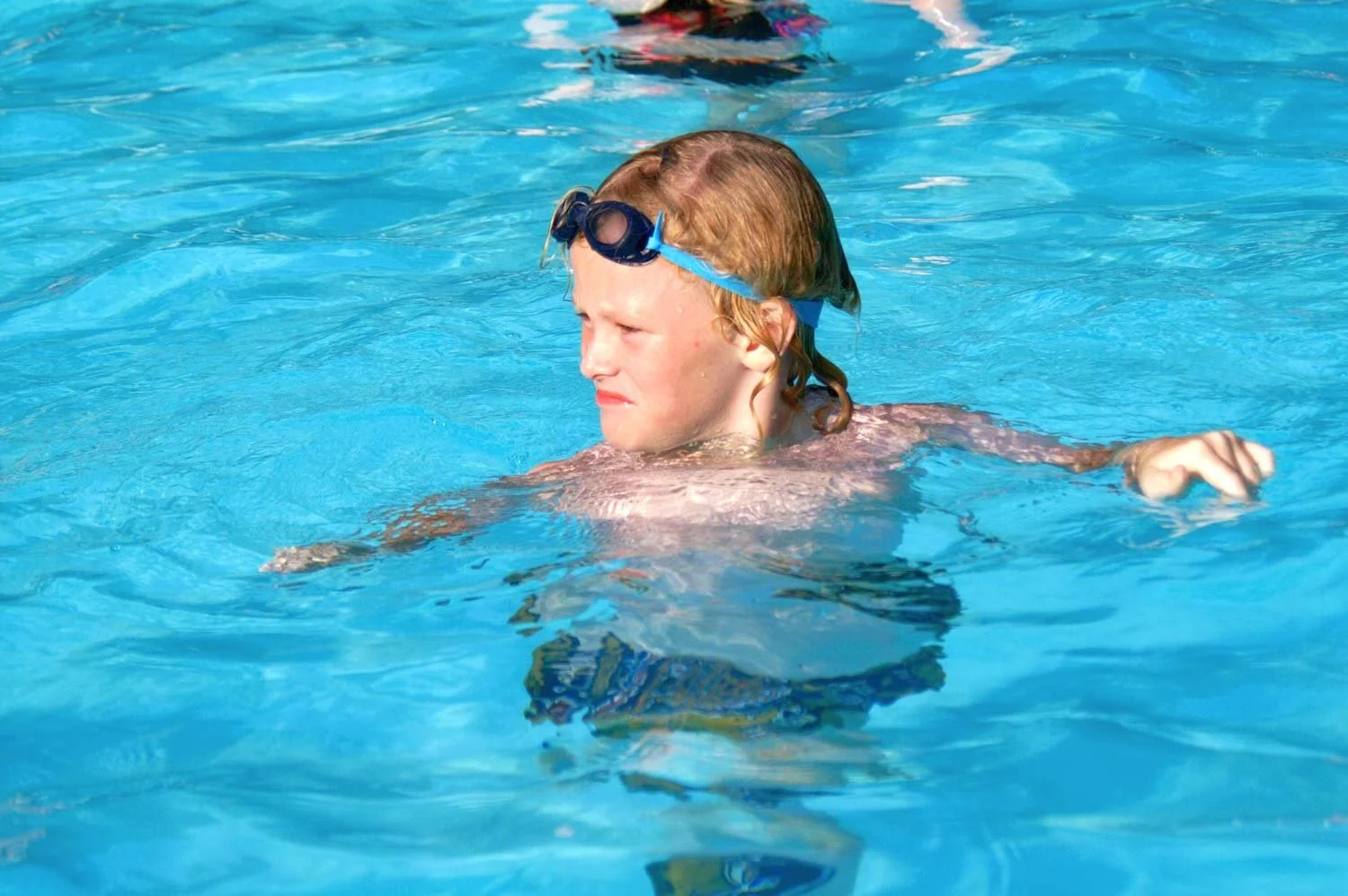 A young boy with red hair swimming in a blue pool, wearing goggles on his forehead and looking focused.