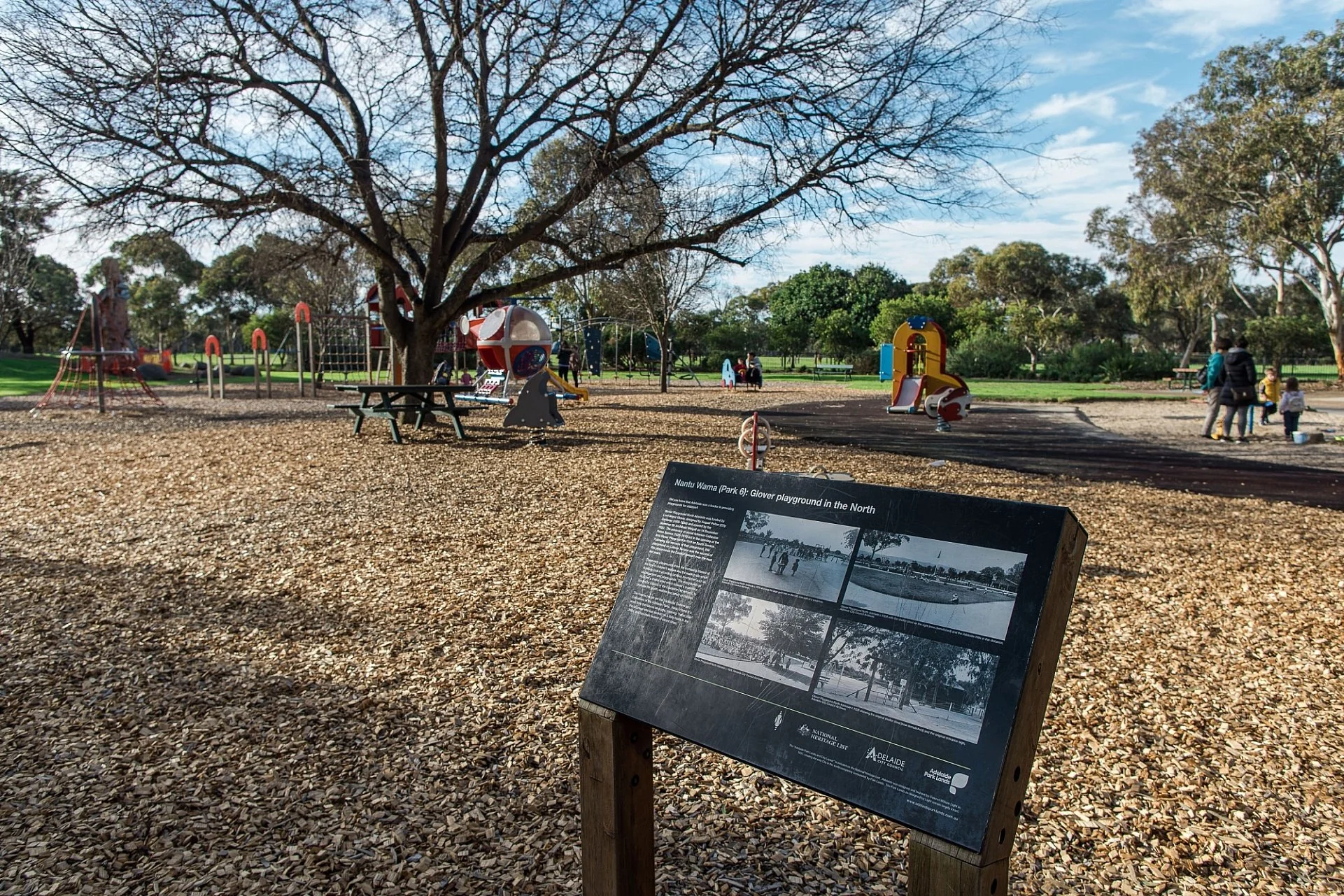 Children playing on colorful playground equipment in a park with trees, benches, and people walking around on a sunny day.