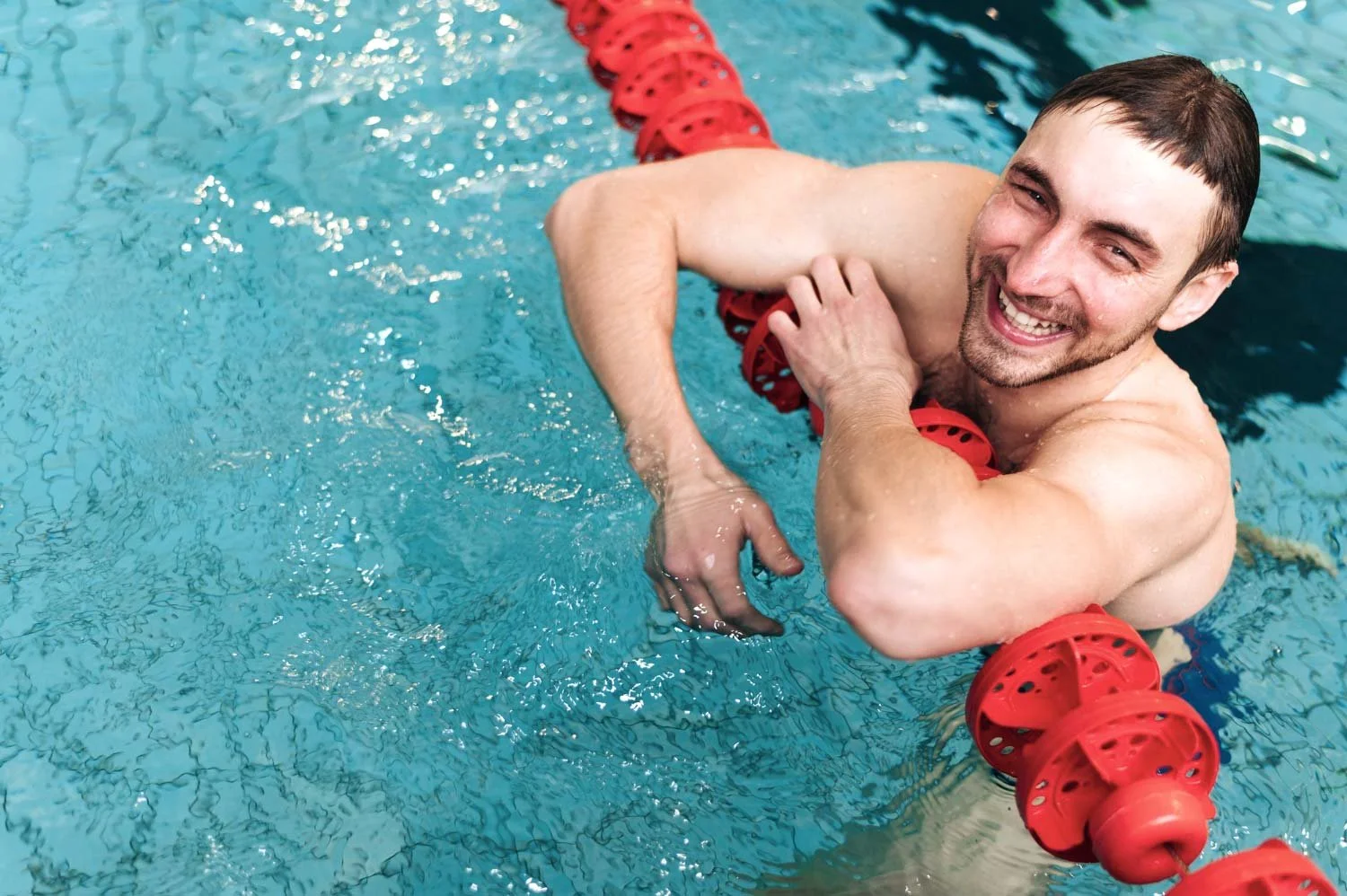 A smiling man swimming in a pool, holding onto a red lane divider.