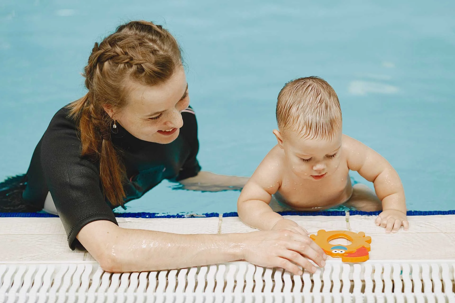 A woman and a baby in a swimming pool playing with a colorful toy near the edge of the pool.