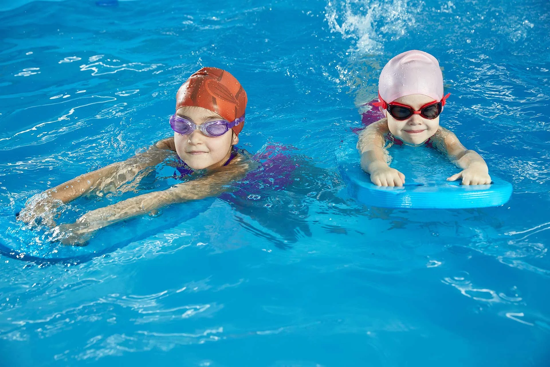 Two young children swimming in a pool, wearing goggles and swim caps, with one using a kickboard for support.