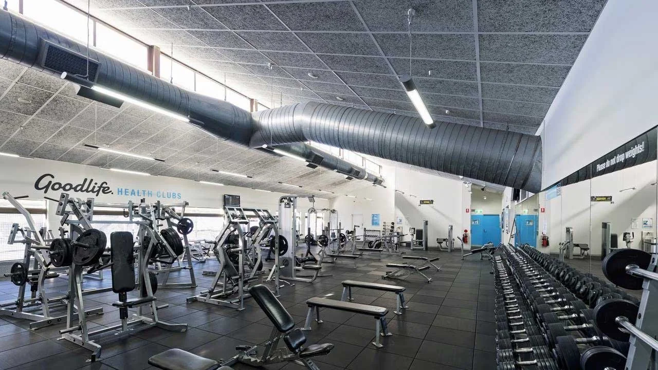 Empty gym with various weightlifting equipment, benches, and Dumbbells, large ventilation duct on ceiling, large mirror on wall, bright lighting, and windows letting in natural light.