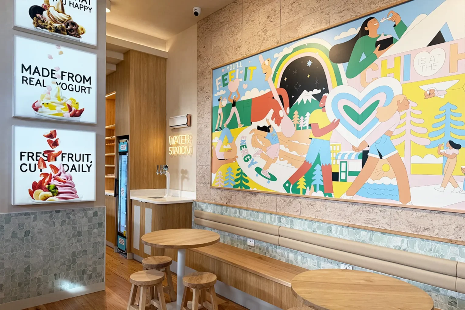 Interior of a cafe with a colorful mural showing people, a rainbow, mountains, and animals, along with ice cream advertisements on illuminated signs.