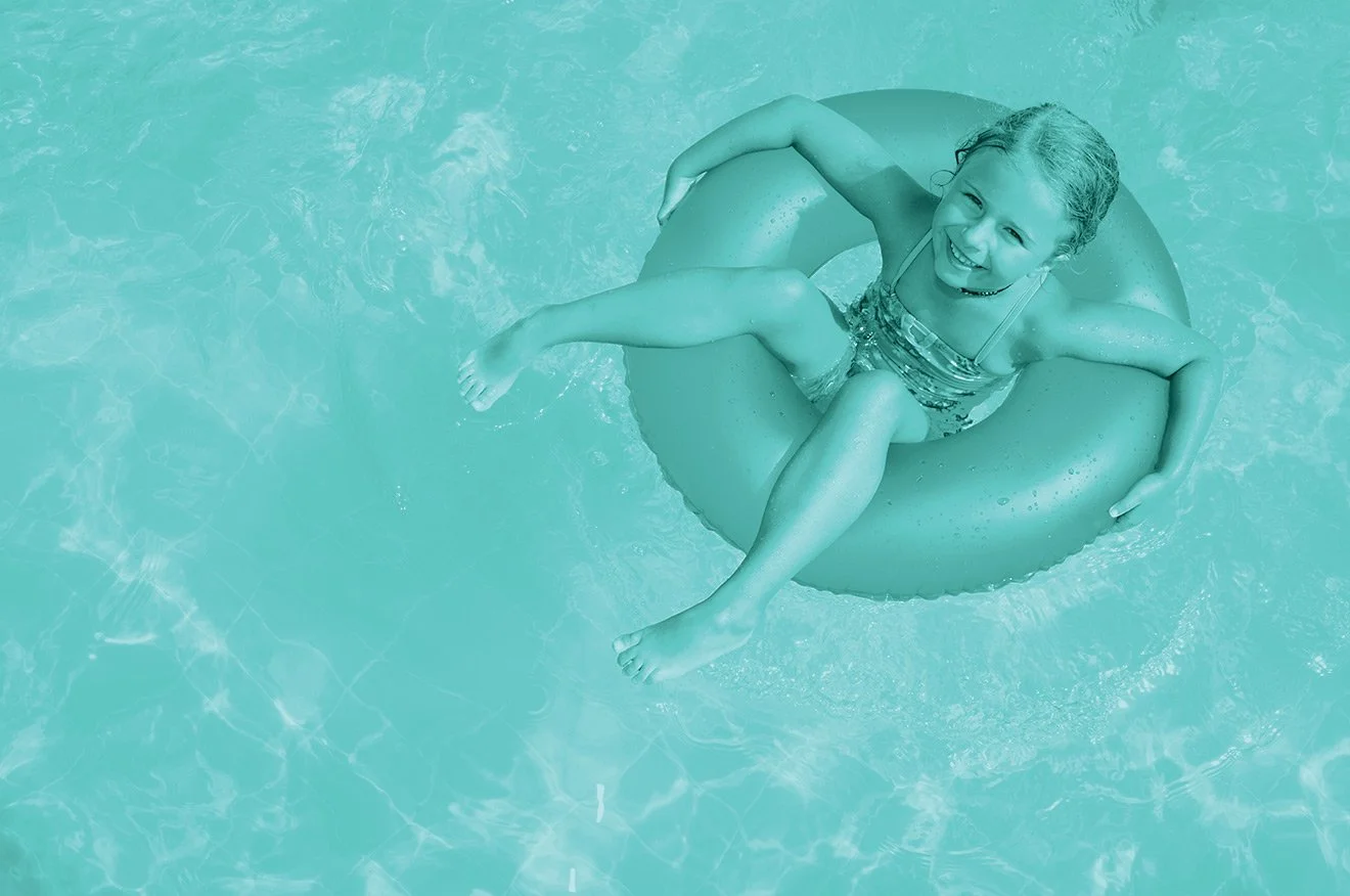 A young girl is sitting on a flotation ring in a swimming pool, smiling and looking up.