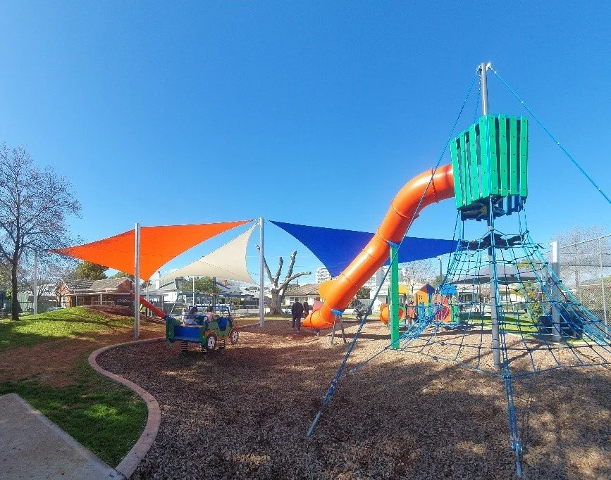 Playground with a large orange slide, a climbing structure, and shade sails in a park under a clear blue sky.