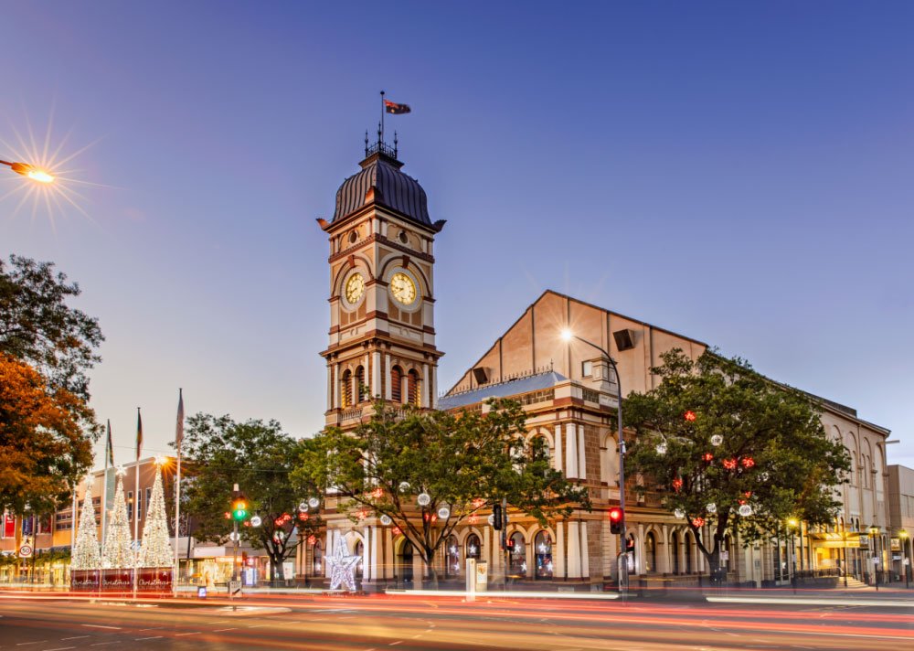 A historic clock tower with a Christmas display, trees, and traffic lights at dusk.