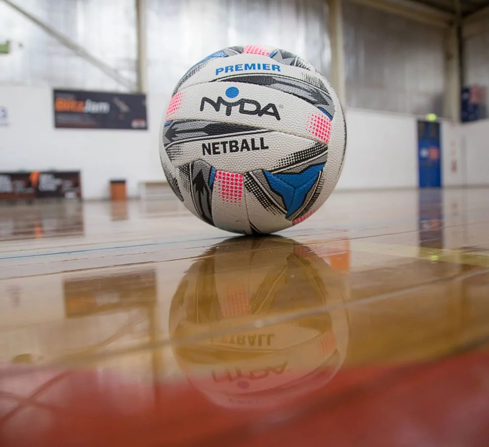 A colorful netball with brand and design markings is on a polished wooden indoor sports court, with a blurred background of the court's walls and signage.