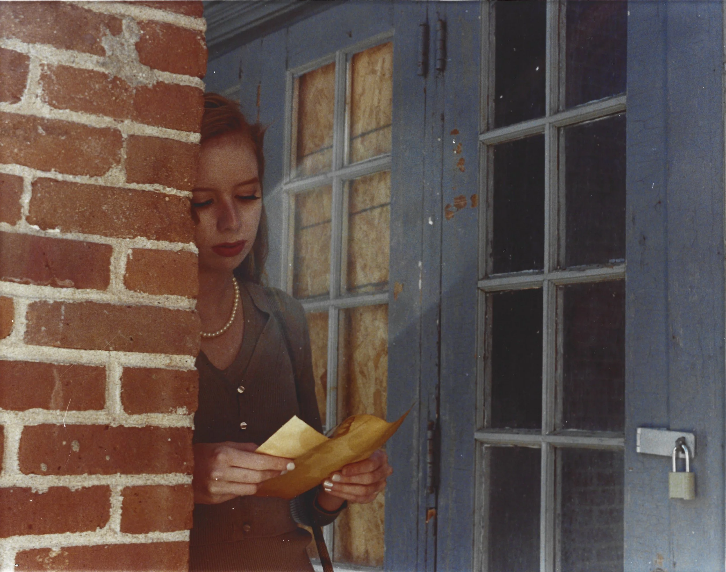 A woman with red hair, wearing a pearl necklace and a gray top, looks down at a yellow envelope she is holding. She is standing near a red brick wall and a blue wooden door with glass panes and a padlock.