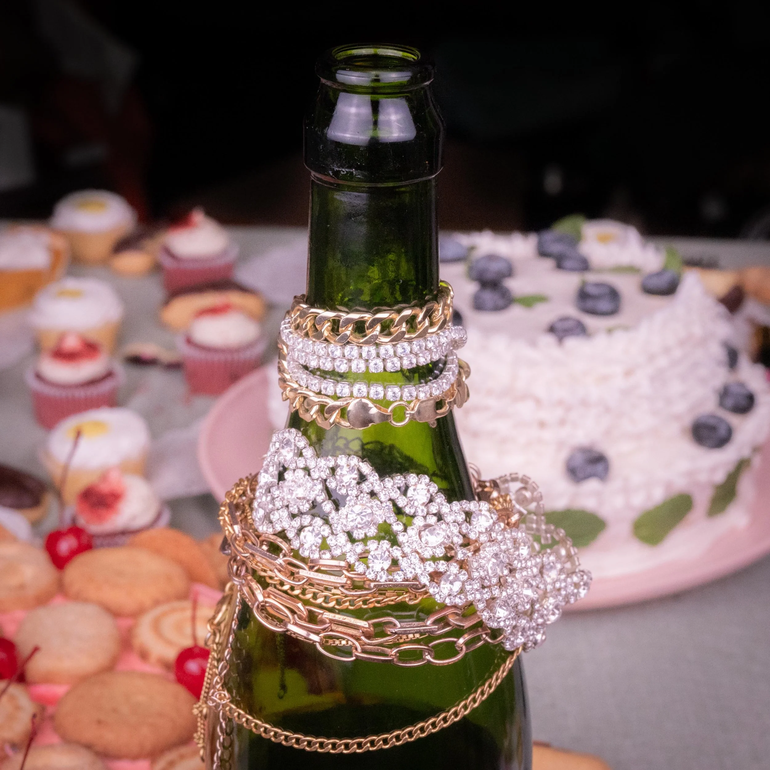 Green glass bottle stacked with various gold and silver jewelry, including rings, bracelets, and chains, with a dessert table featuring cupcakes and a blueberry cake in the background.