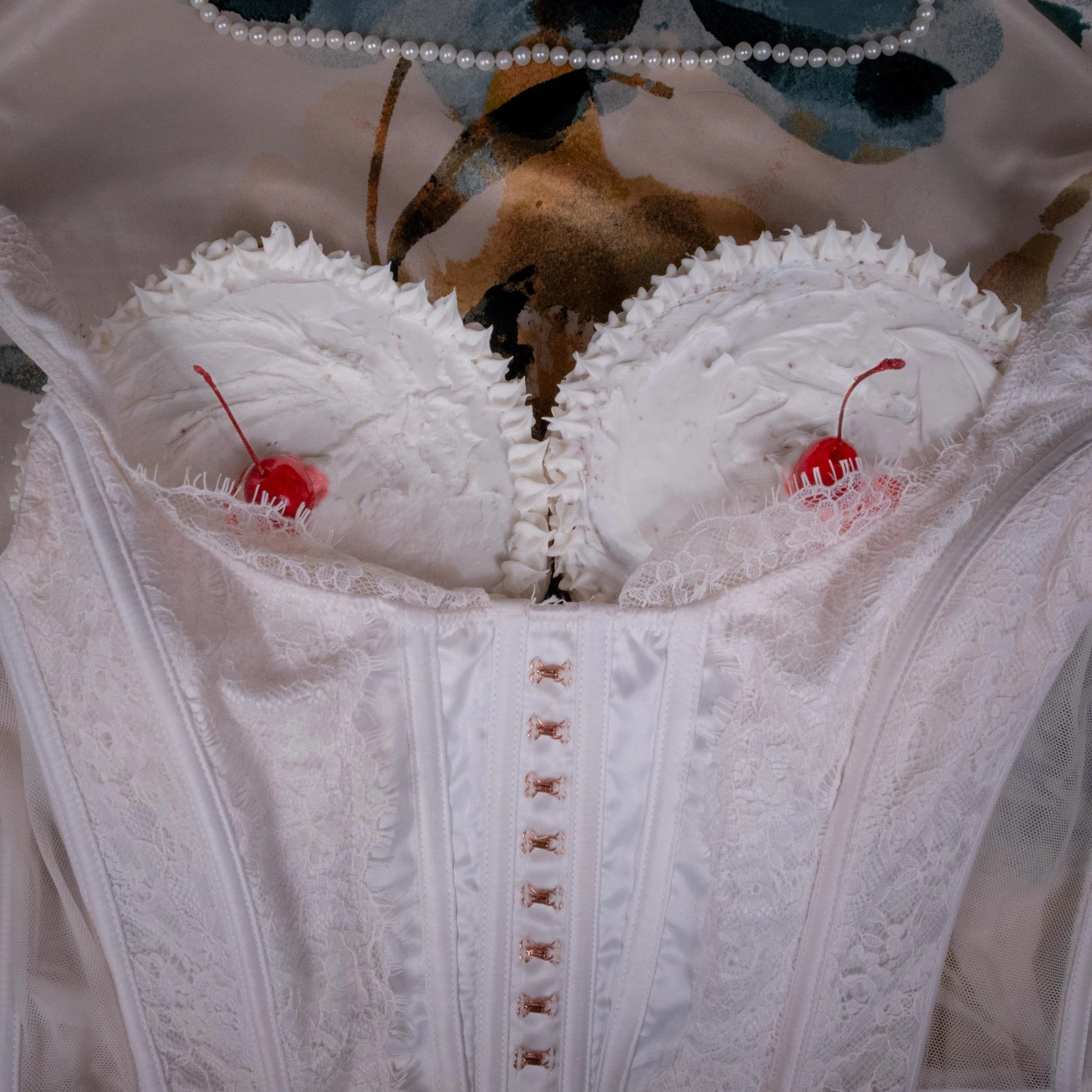 A white wedding cake with two tiers, decorated with white whipped cream and topped with cherries, wrapped in white lace and satin rap. Pearl necklace is draped above the cake.