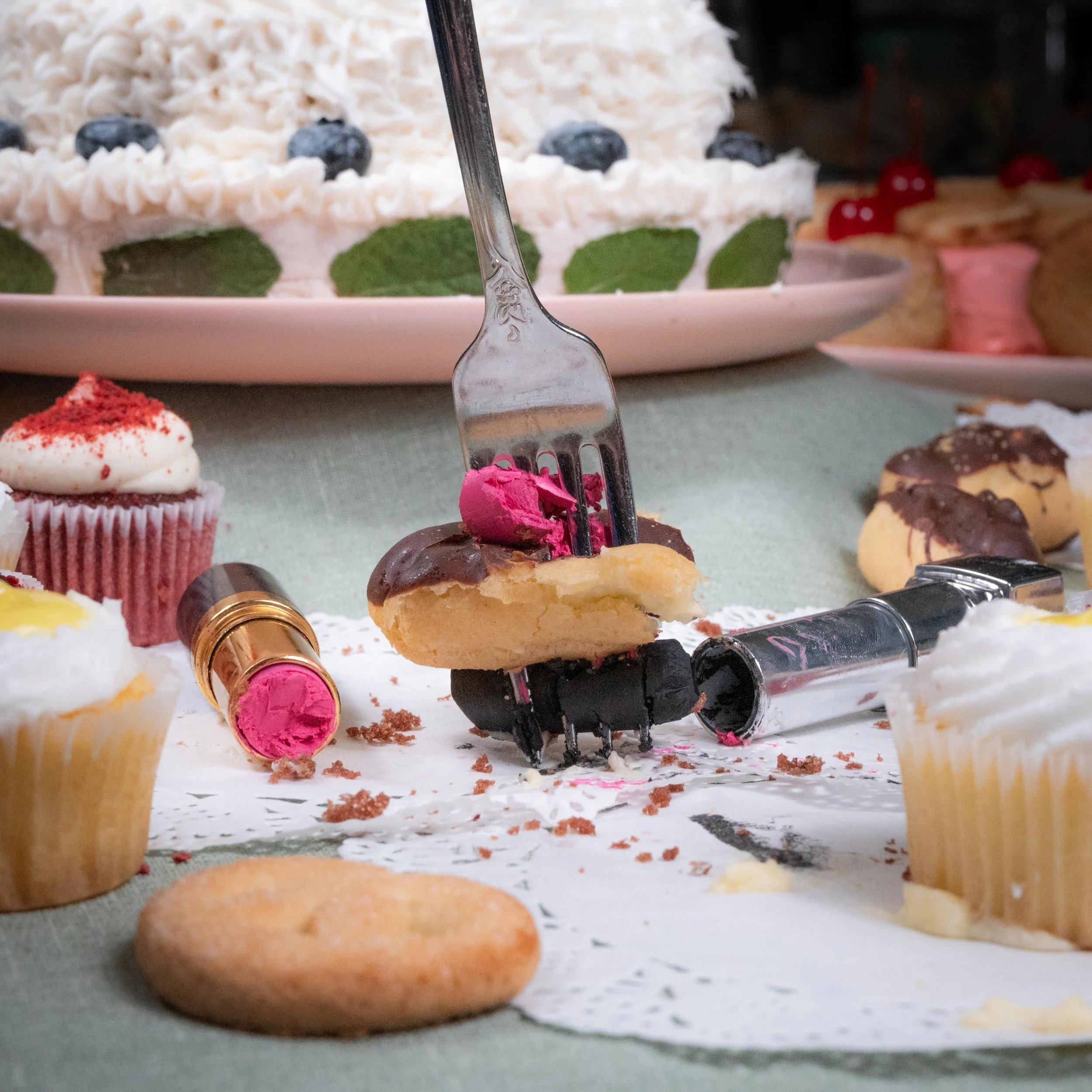 Dirty dessert table with crumbs, lipstick, lipstick case, cupcakes, and a partially eaten cake in the background