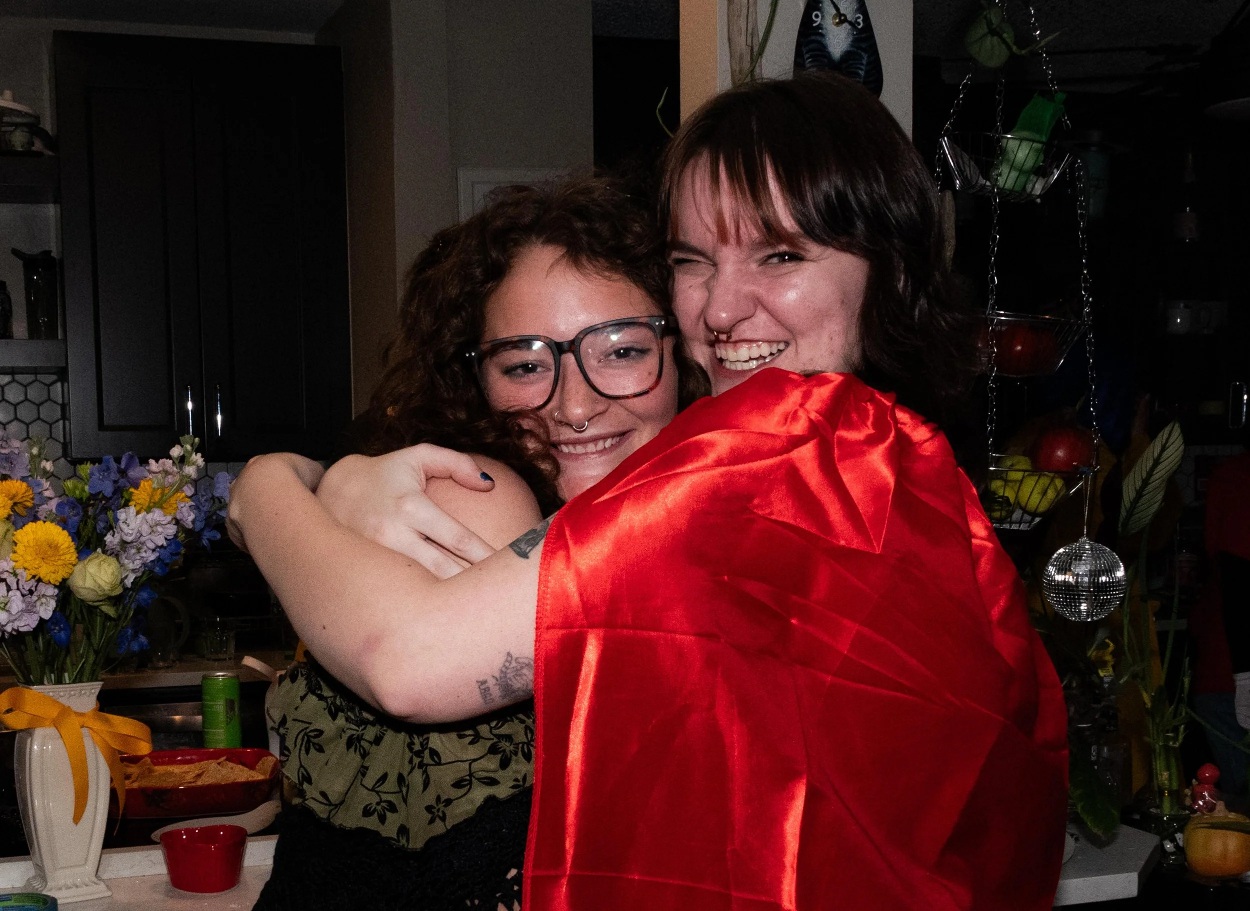 Two women hugging and smiling, one wearing glasses and a floral top, the other with shoulder-length dark hair and a septum piercing, in a kitchen decorated with flowers and hanging fruit baskets.