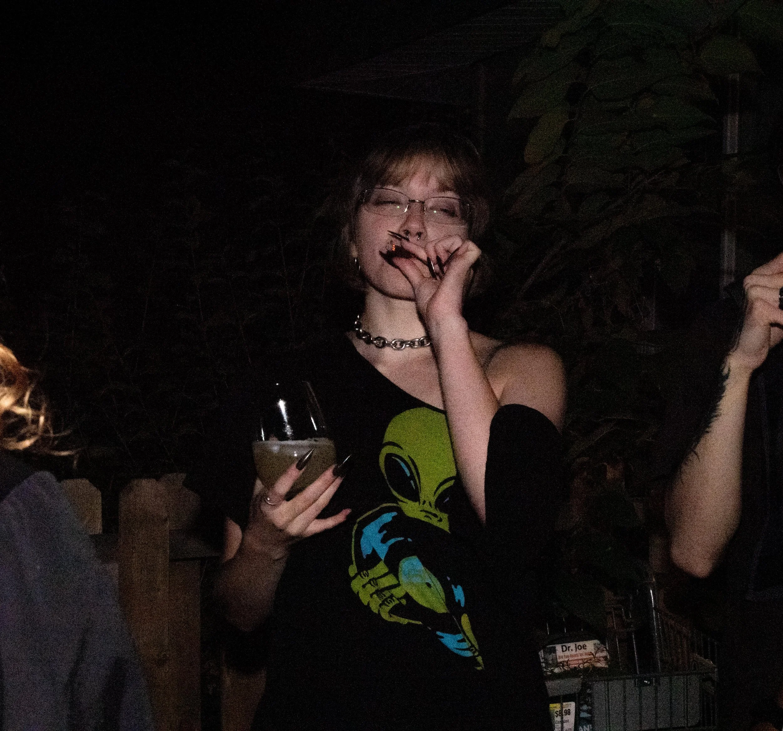 Young woman with glasses and short hair, wearing a black shirt with a green alien graphic, holding a drink in her left hand, and making a gesture near her mouth with her right hand at night.