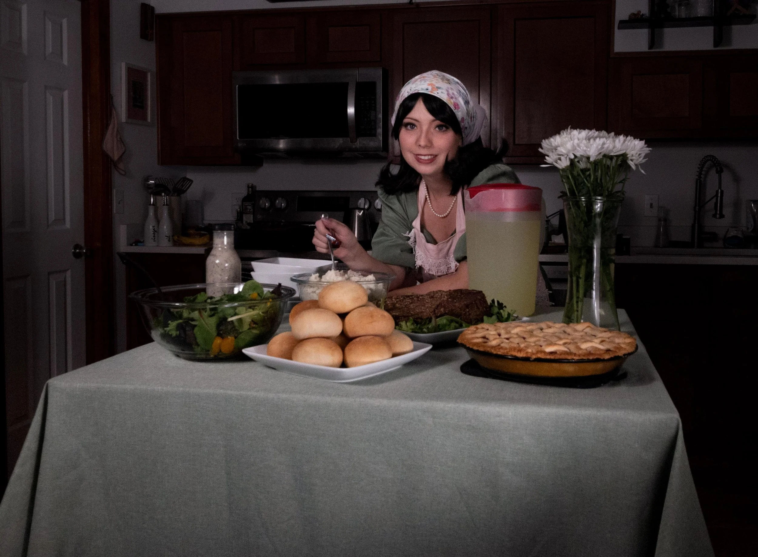 A woman in a kitchen preparing food at a table with various dishes and flowers.