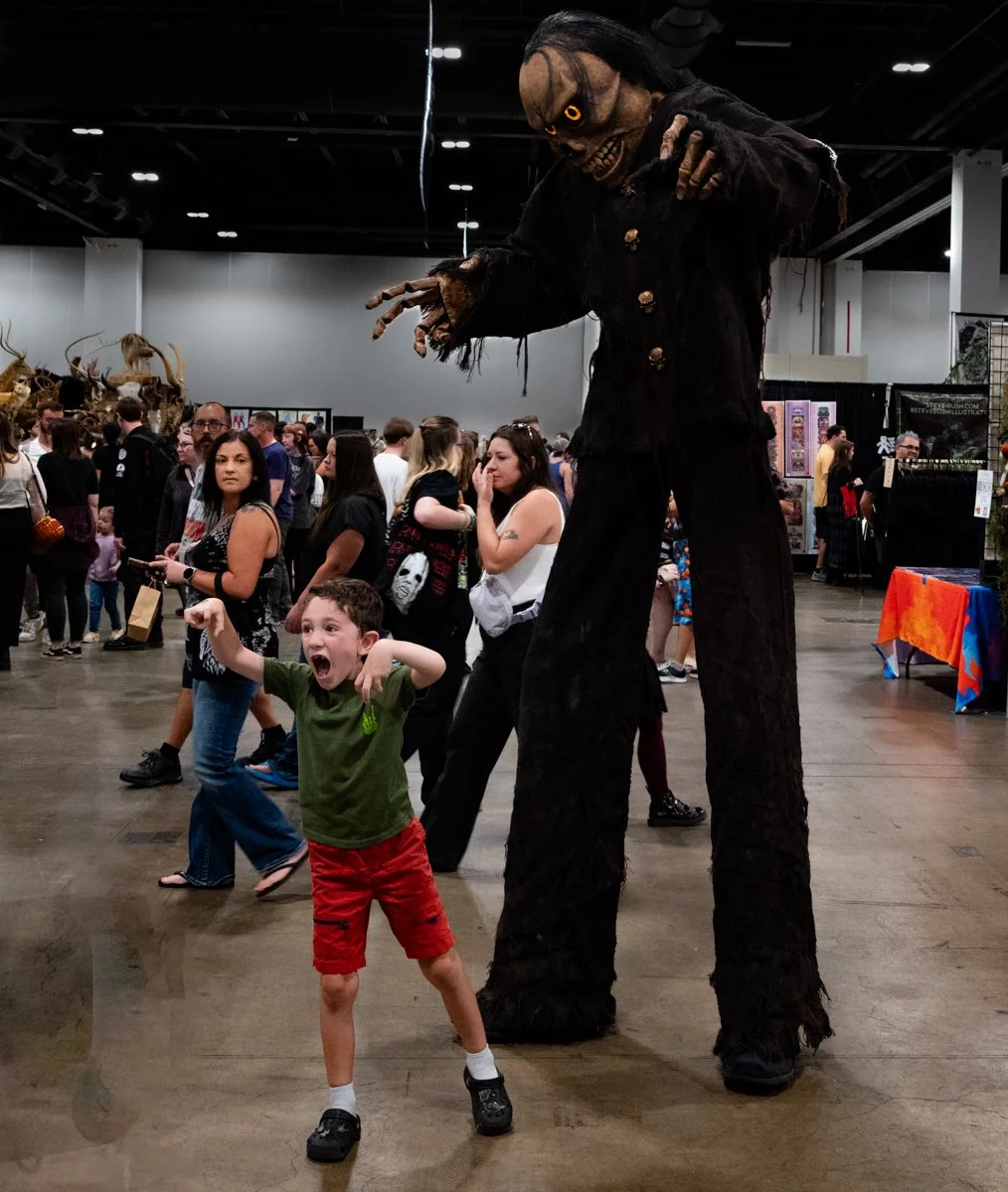 A person in a large costume of a monster with long arms, face, and black clothing standing over children and adults at a convention or event hall.