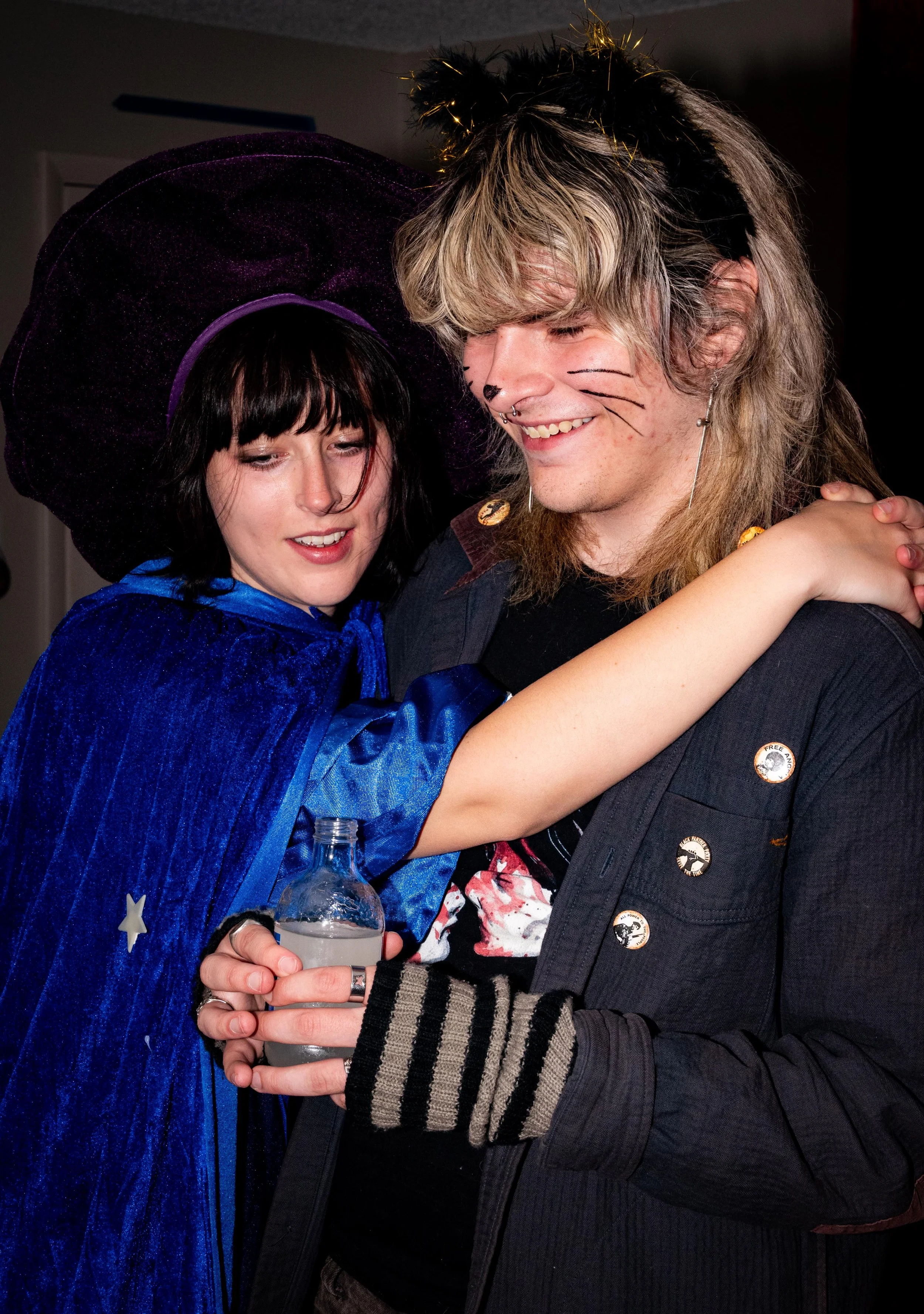 Two people embracing, dressed in costumes, smiling, with one wearing pointy hat and a cape, and the other with late 80s/early 90s style hair, with cat face makeup and black jacket with pins, holding a bottle of water.