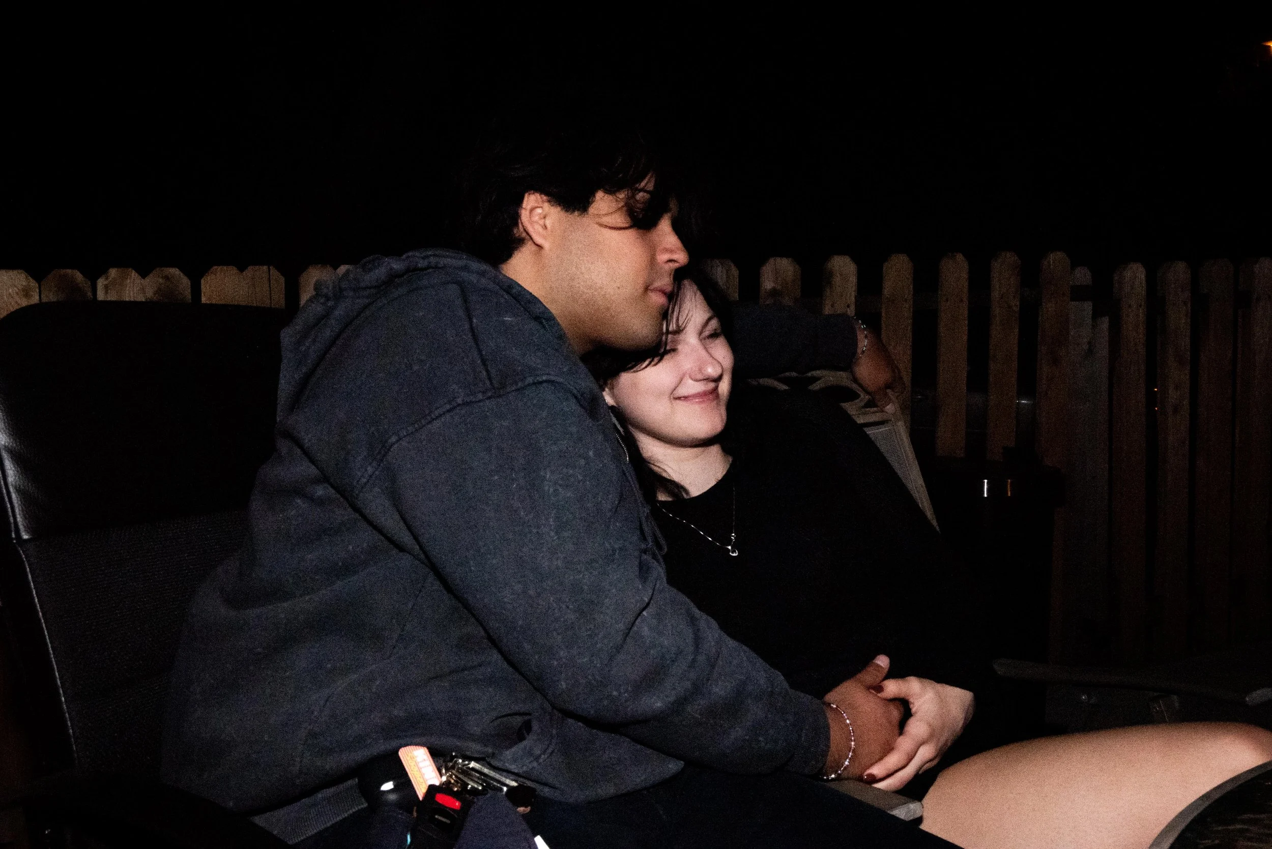 A young couple sitting close together on outdoor chairs at night, with the man resting his head on the woman's and both smiling, in front of a wooden fence.