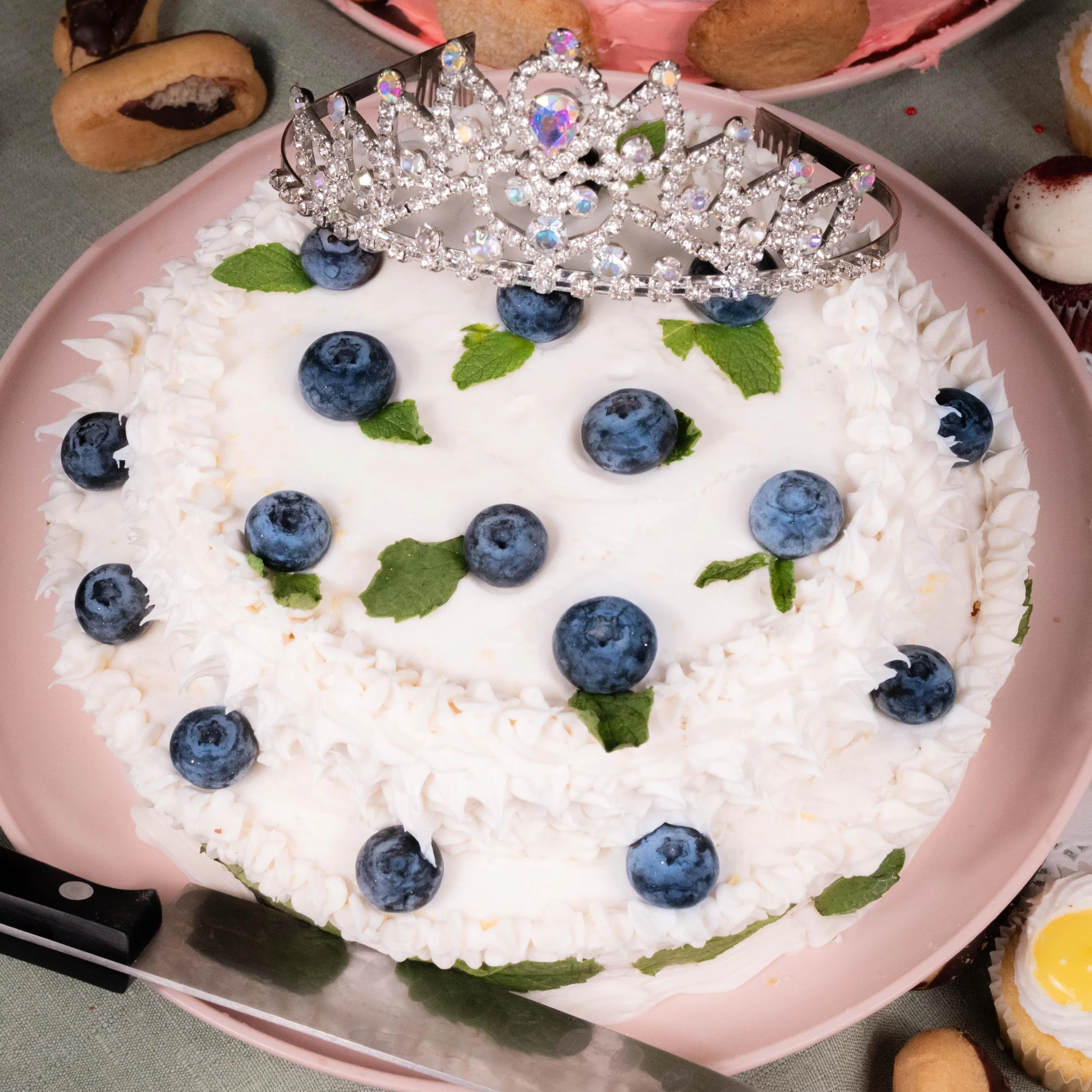 A round birthday cake decorated with blueberries, mint leaves, and whipped cream, topped with a tiara-shaped decoration.