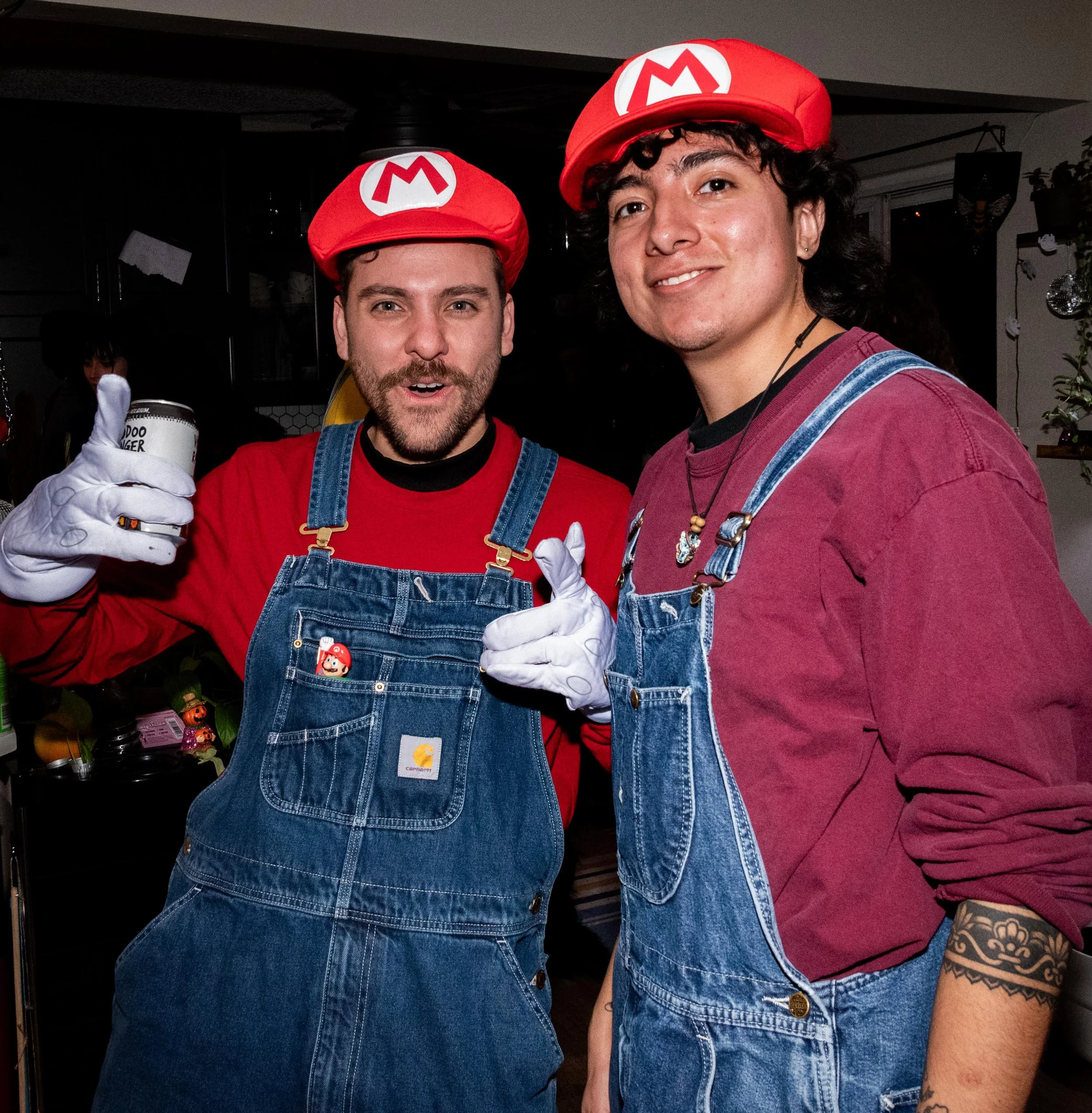 Two men dressed as Mario and Luigi from Nintendo, wearing red hats with 'M' and 'L' logos, denim overalls, and white gloves, celebrating at a party with smiles and pointing at the camera.