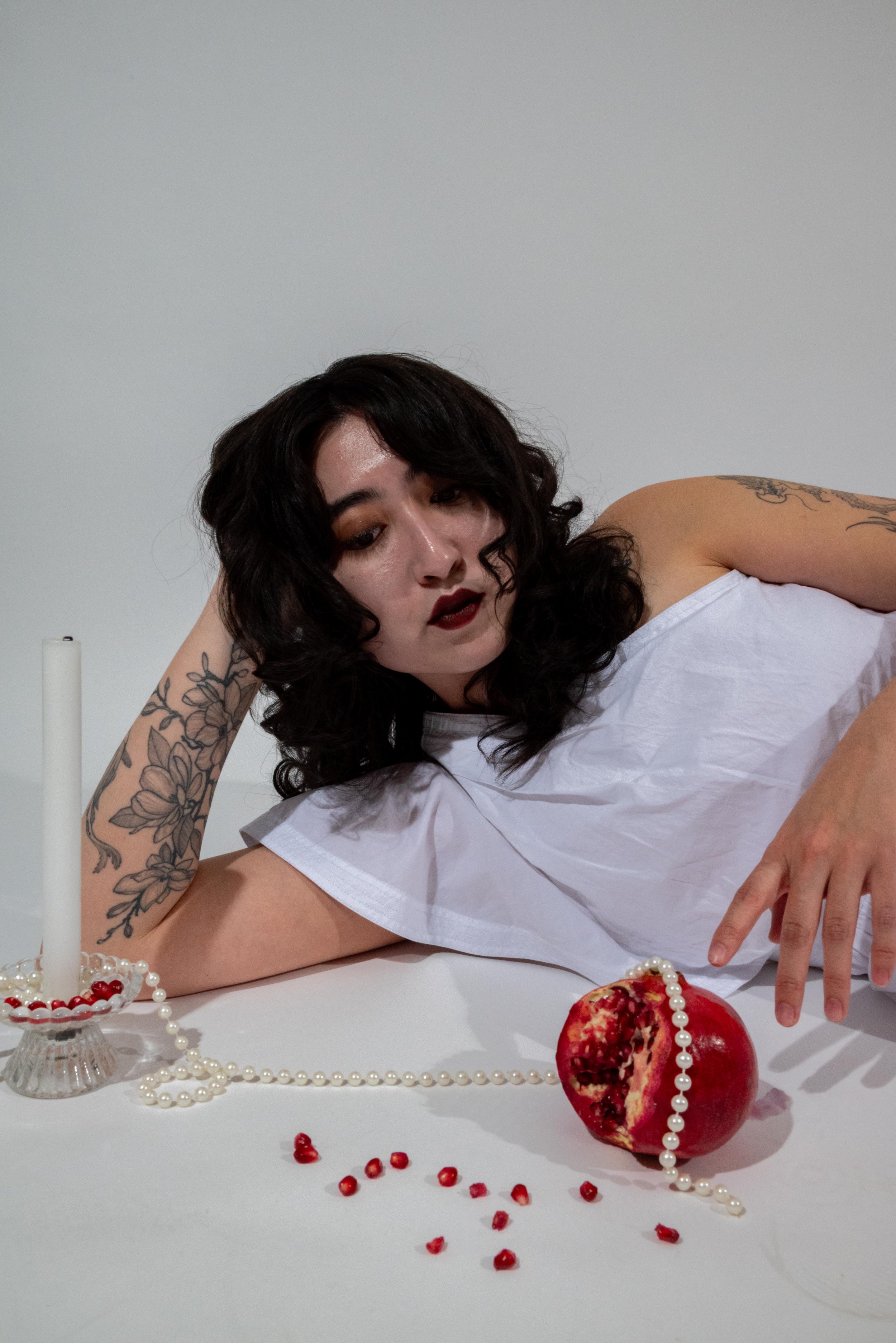 A woman with dark curly hair and tattoos lying on a white surface, wearing a white top, surrounded by a pomegranate, pearl necklace, and a candlestick with a candle.