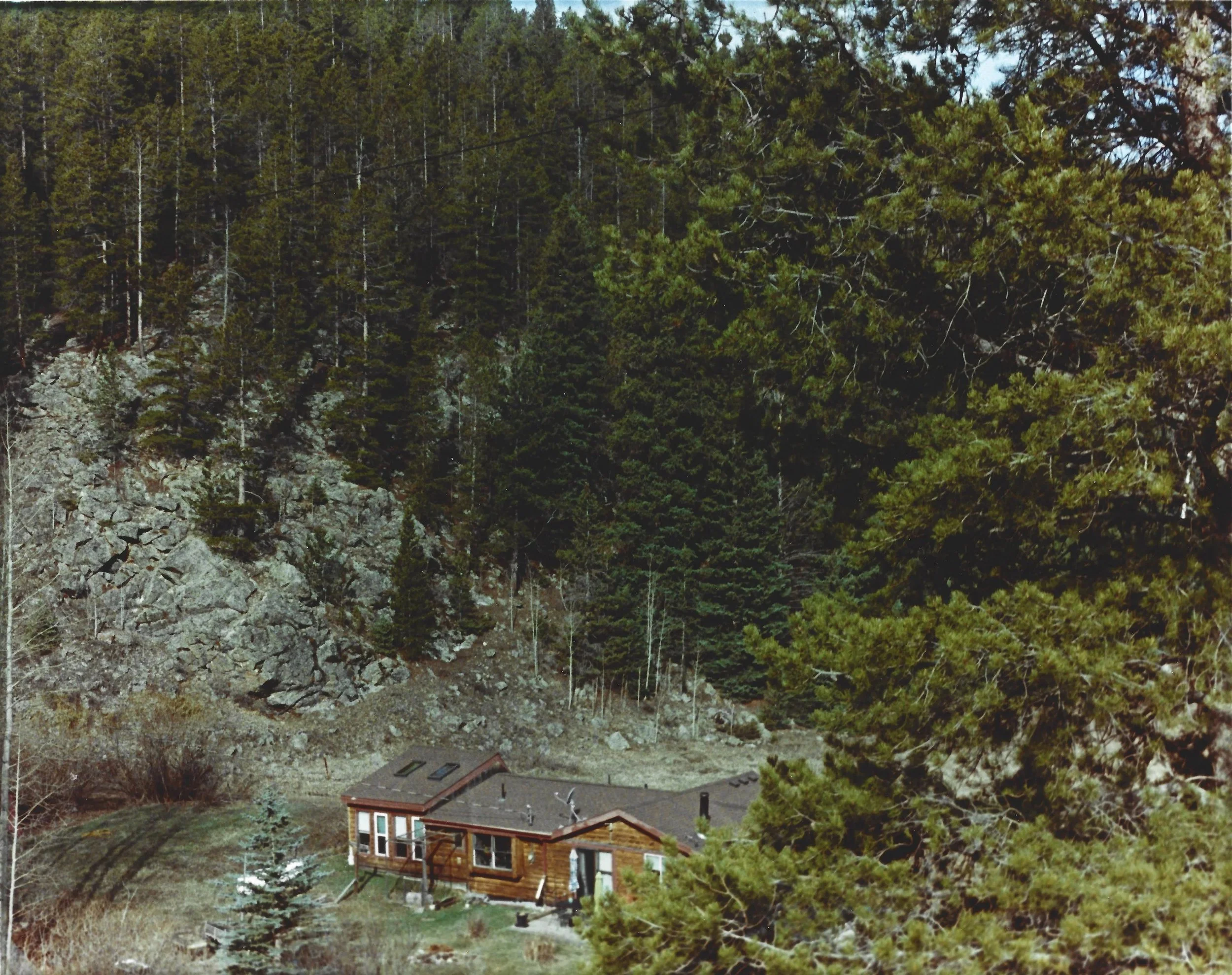 A small wooden house in a wooded area with tall trees and rocky terrain surrounding it.