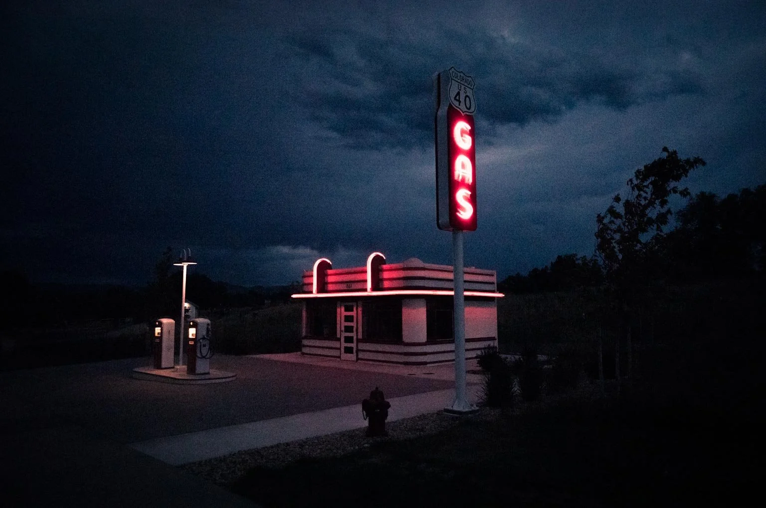 A vintage gas station with a neon sign spelling 'GAS' on a tall pole, illuminated in red against a dark, cloudy sky, with two gas pumps nearby.