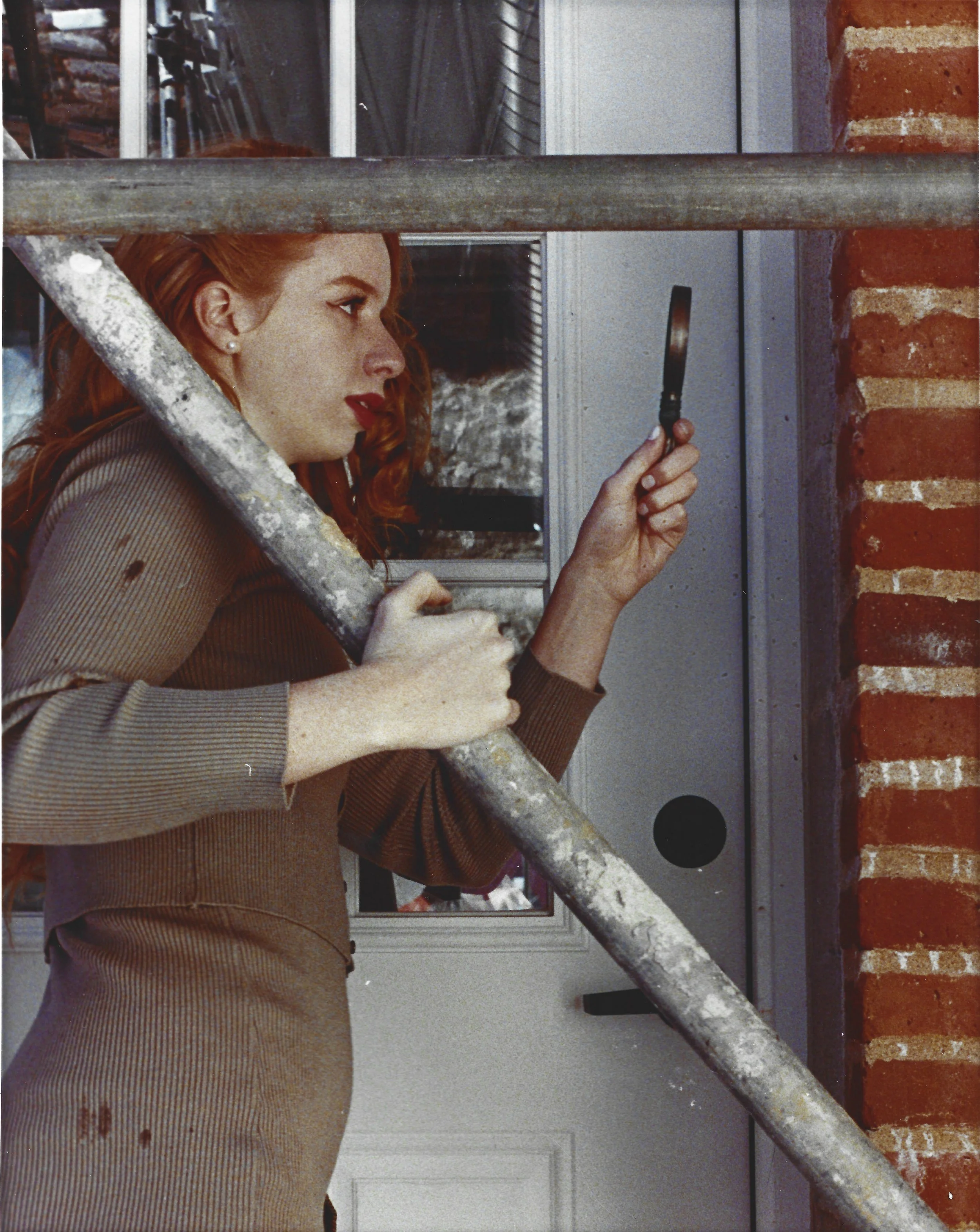 A woman with red hair and red lipstick, wearing a brown dress, stands on a staircase and looks through a magnifying glass at a door or panel.