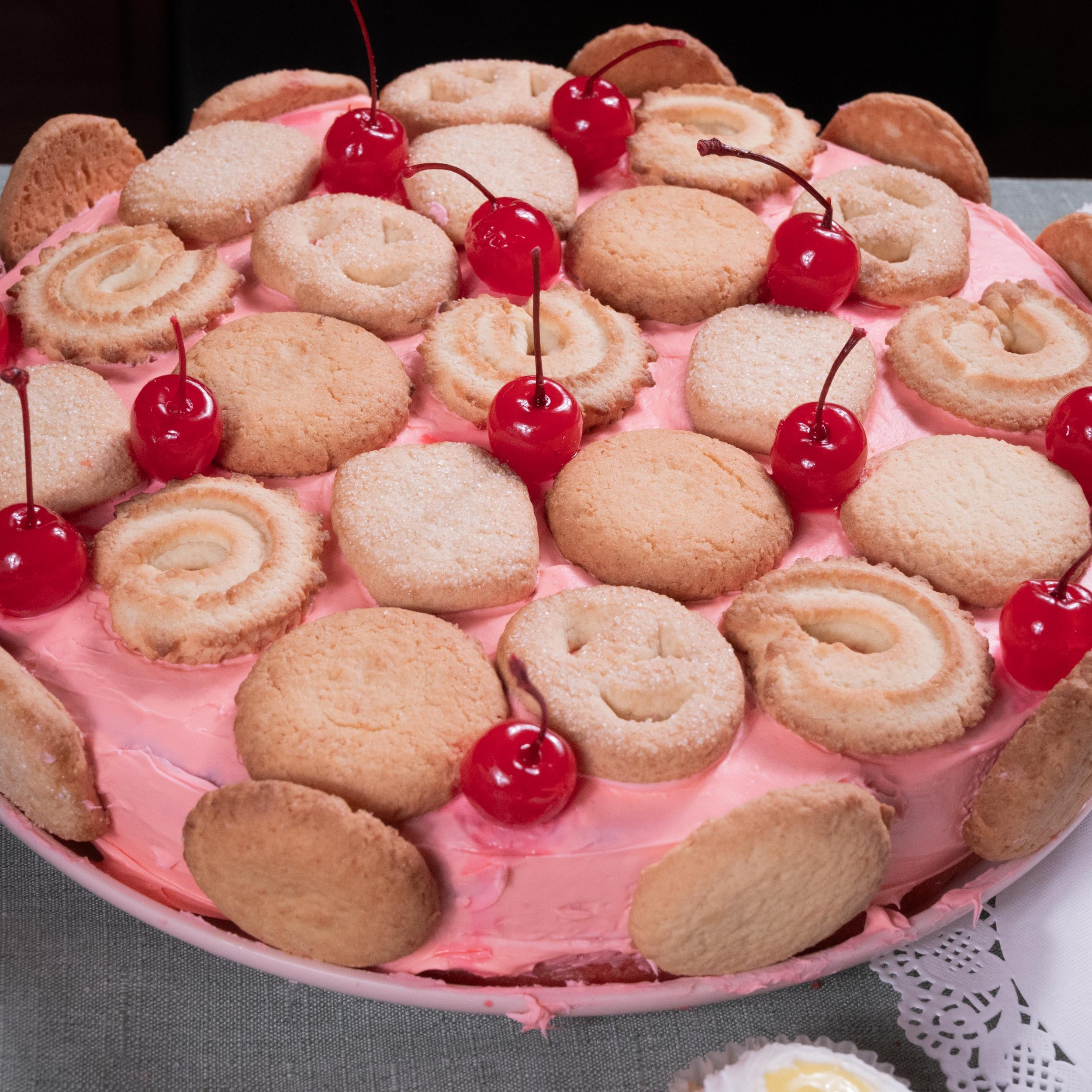 Layered pink frosted cake decorated with smiley face cookies, cherry toppings, and surrounded by additional cookies.
