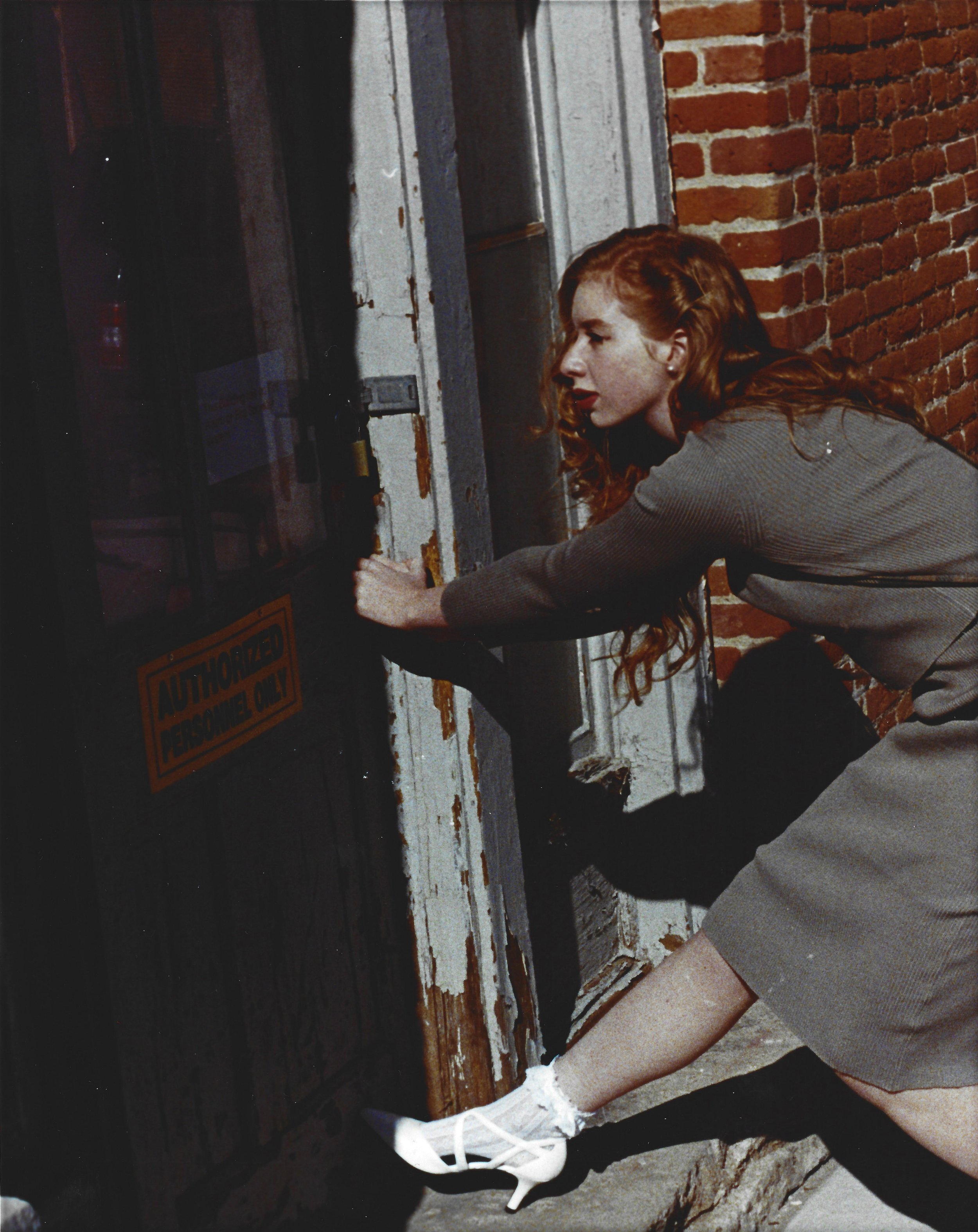 Red-haired woman in a skirt and high-heeled shoes attempting to force open a door with a lock, standing outside a brick building.
