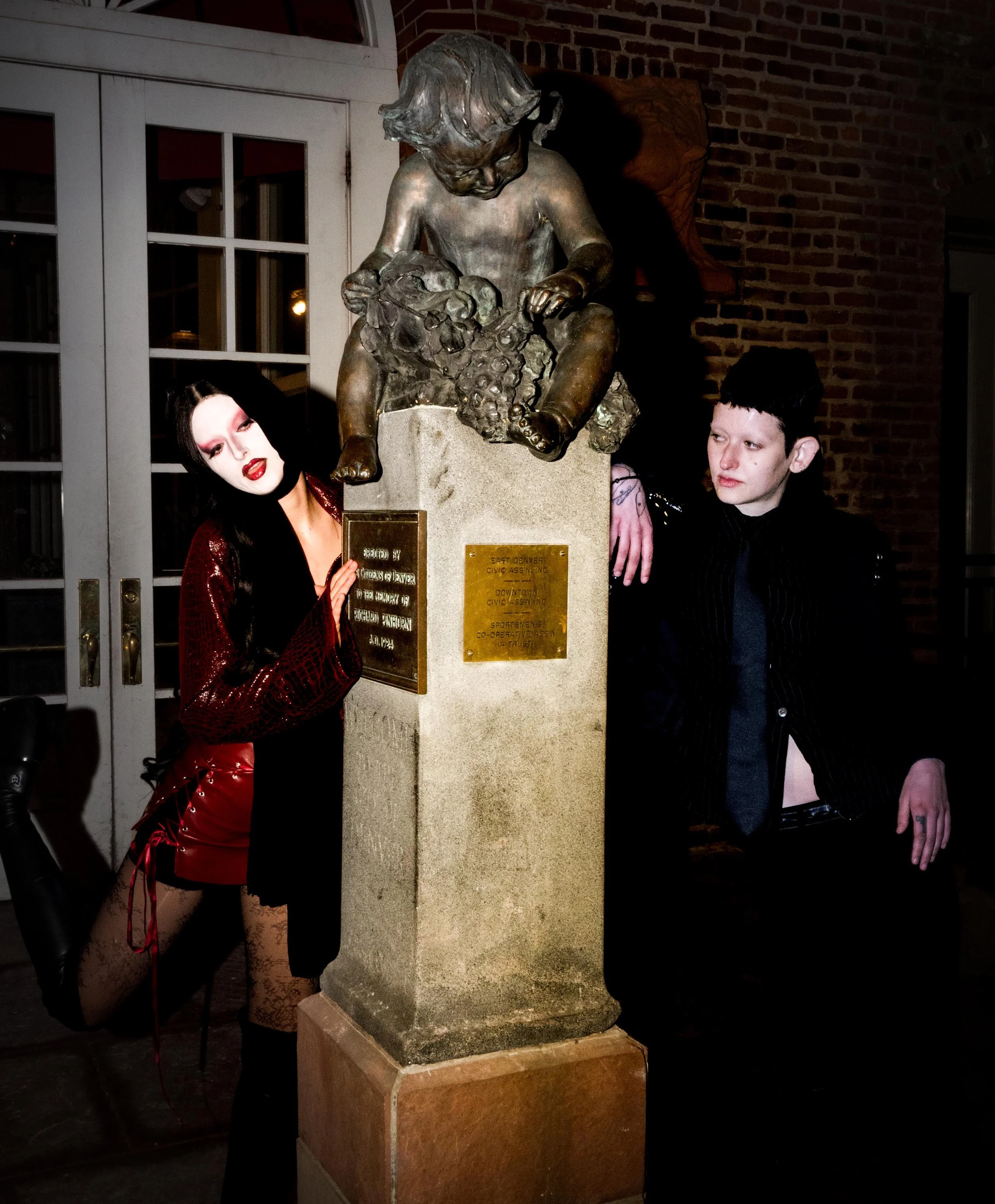 Two individuals dressed in goth style clothing stand beside a statue of a boy holding a bunch of grapes. The woman, on the left, has long black hair, pale makeup, and wears a red and black outfit with lace stockings. The man, on the right, has short black hair, pale skin, and wears a black pinstripe suit. The statue is mounted on a stone pedestal with plaques, situated outside near brick walls and glass doors.