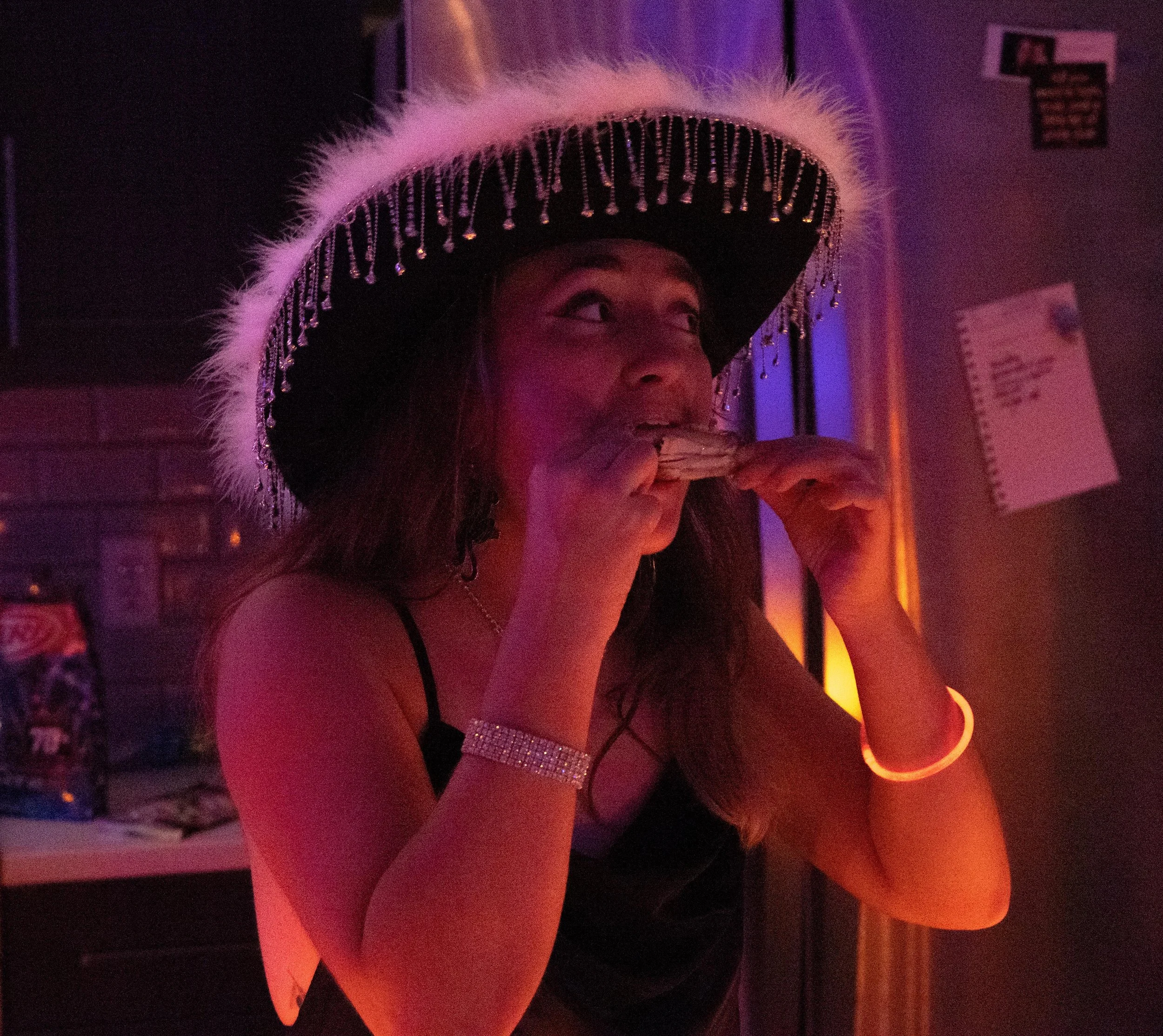 A woman wearing a large, decorated hat with white feathers and hanging beads, holding and biting into a slice of pizza in a dimly lit setting with colorful lights.