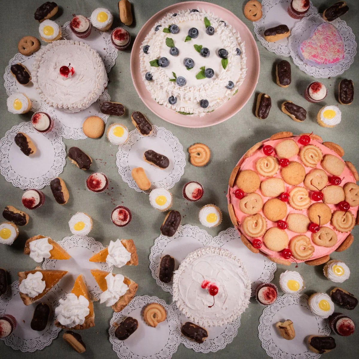 Assorted cakes and cookies on a table, including a cake with blueberries and mint leaves, a pink cake with mini cookies and cherries, and various cupcakes, cookies, and chocolate-covered pastries arranged on paper doilies.