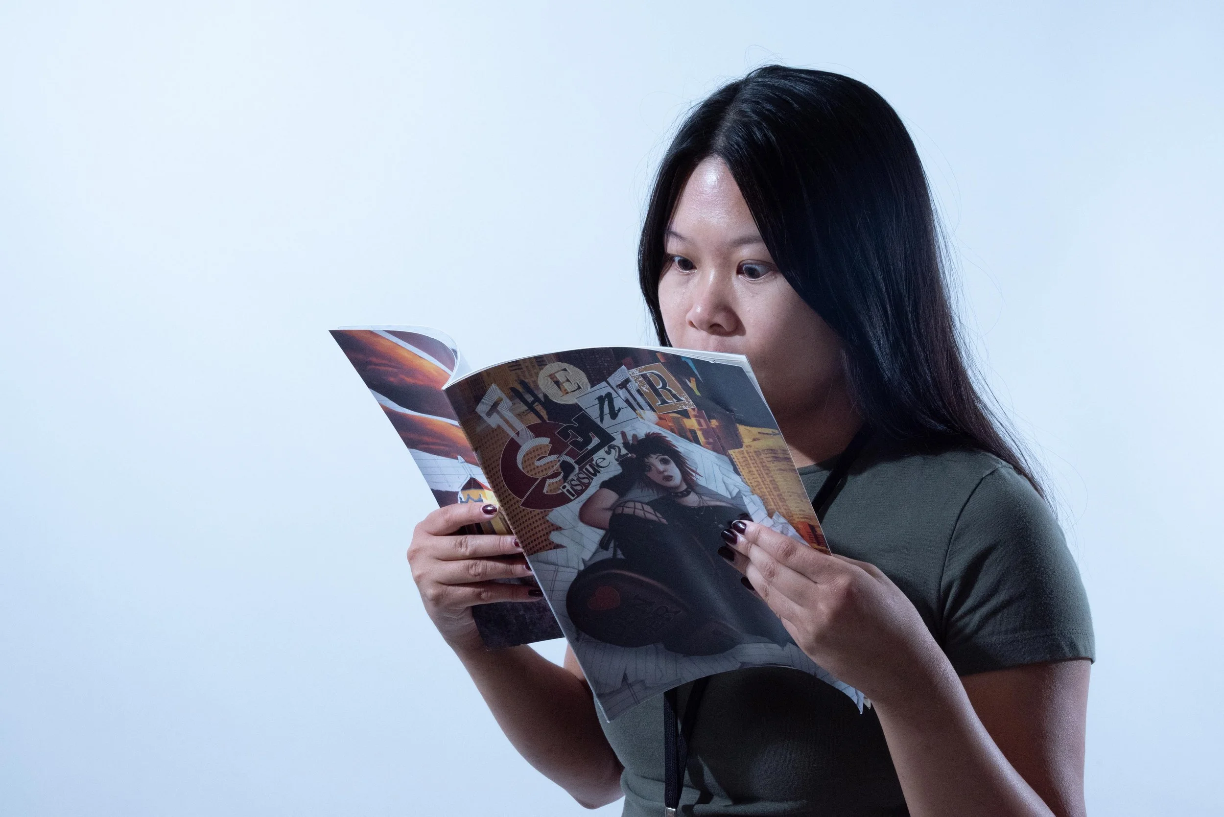 A woman with black hair reads a magazine titled 'The Issue', featuring a black-haired woman in punk attire on the cover, against a plain light background.