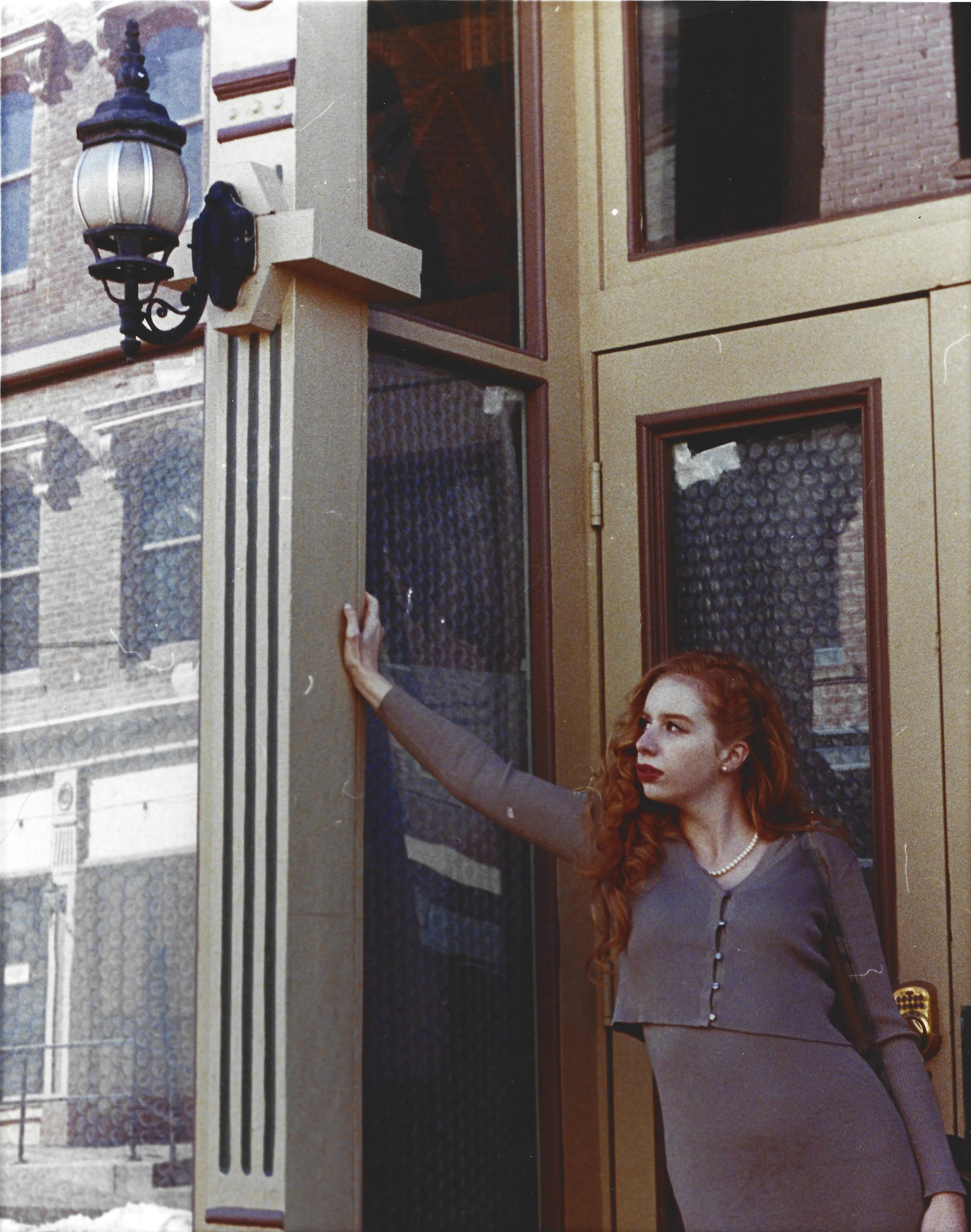 A woman with long, curly red hair standing at a glass door, holding the door handle with her left hand, wearing a gray dress and a pearl necklace. The scene is outdoors in an urban area with brick buildings and an ornate streetlamp overhead.