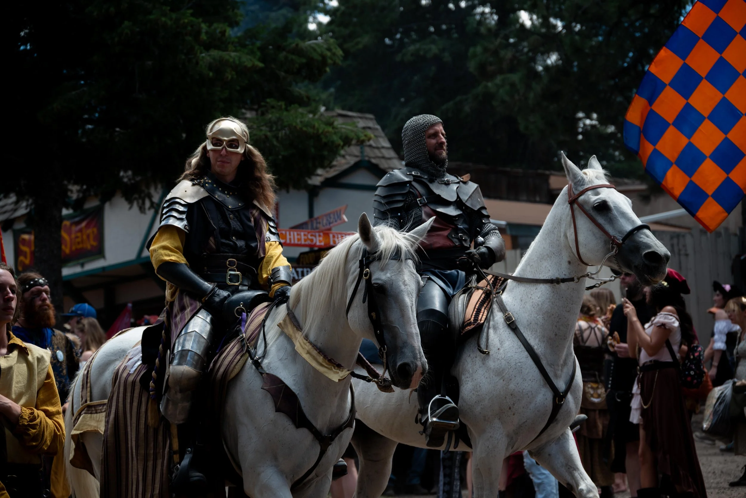 Two people dressed as medieval knights riding white horses at a festival or reenactment event, with a crowd and vendor booths in the background.