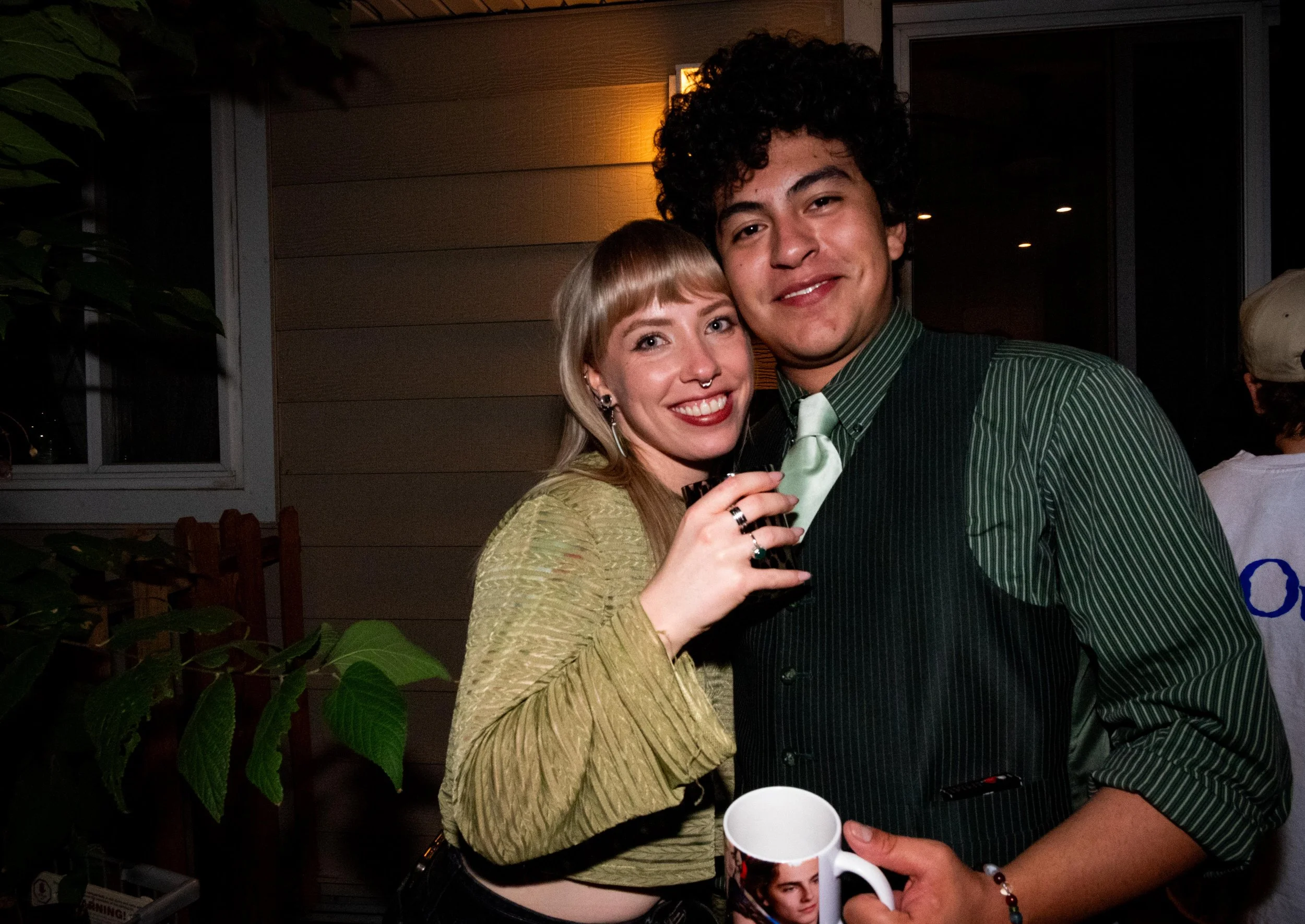 A young woman with blonde hair, dark lipstick, and piercings, smiling and holding a drink, standing next to a young man with curly hair, smiling, dressed in a green shirt and apron, also holding a drink, at an outdoor gathering during evening.
