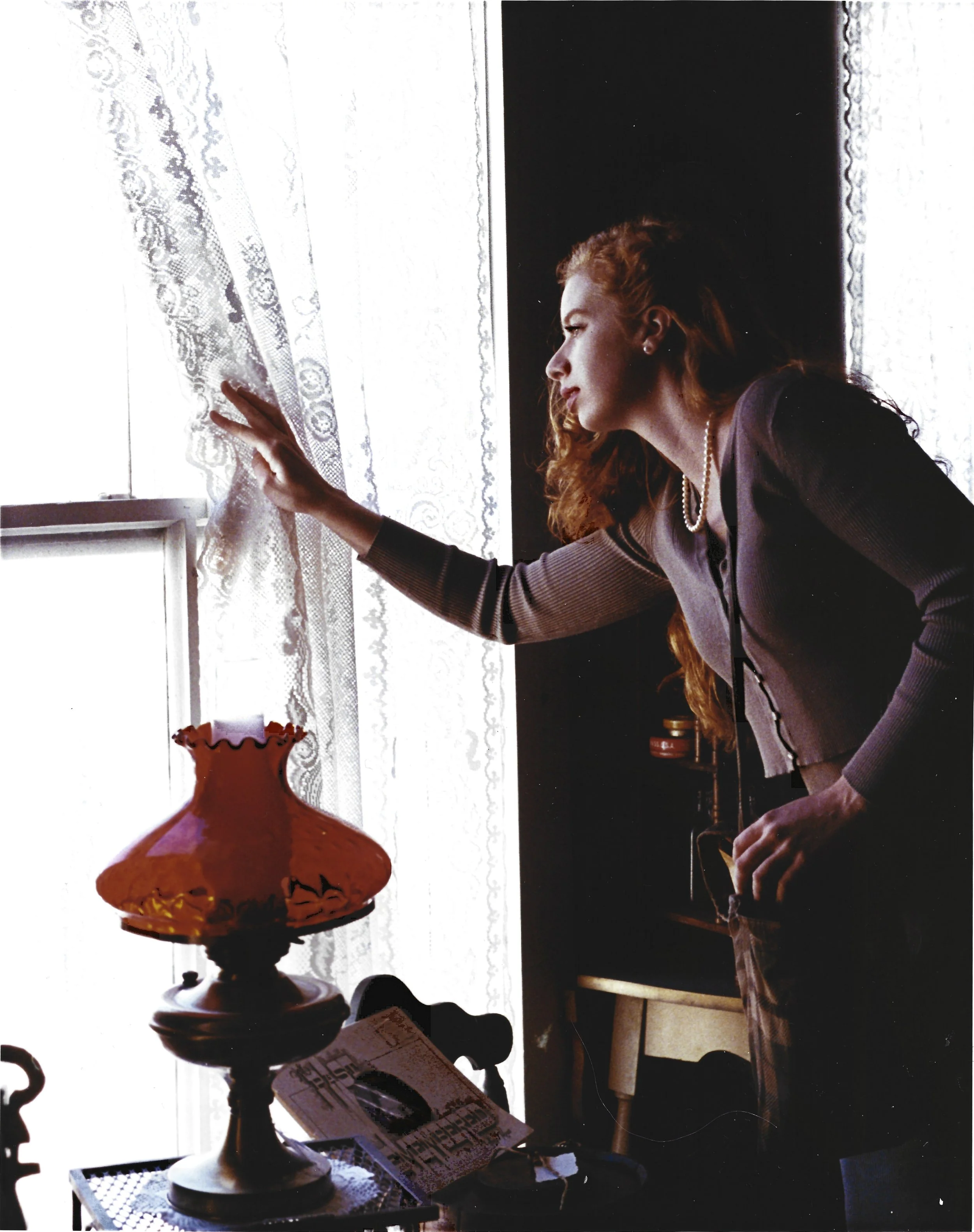 A woman with red hair in a gray long-sleeve top adjusts lace curtains in a sunlit room.