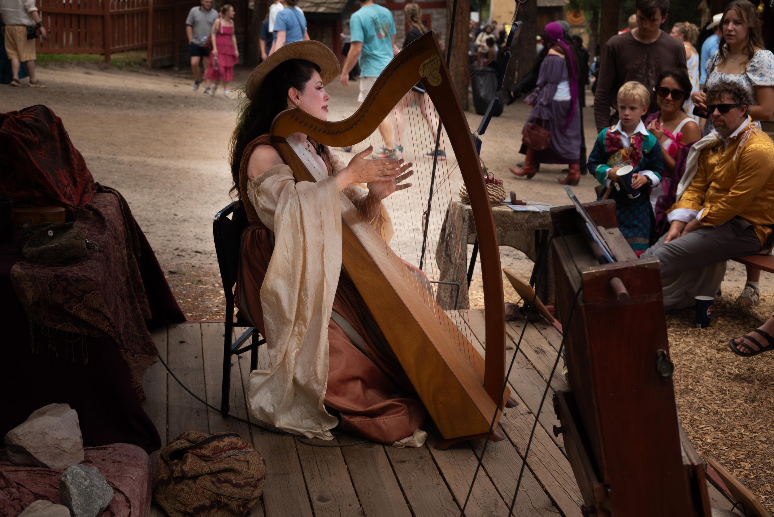 A woman in historical clothing is playing a harpsichord at an outdoor event, with a crowd of people watching.