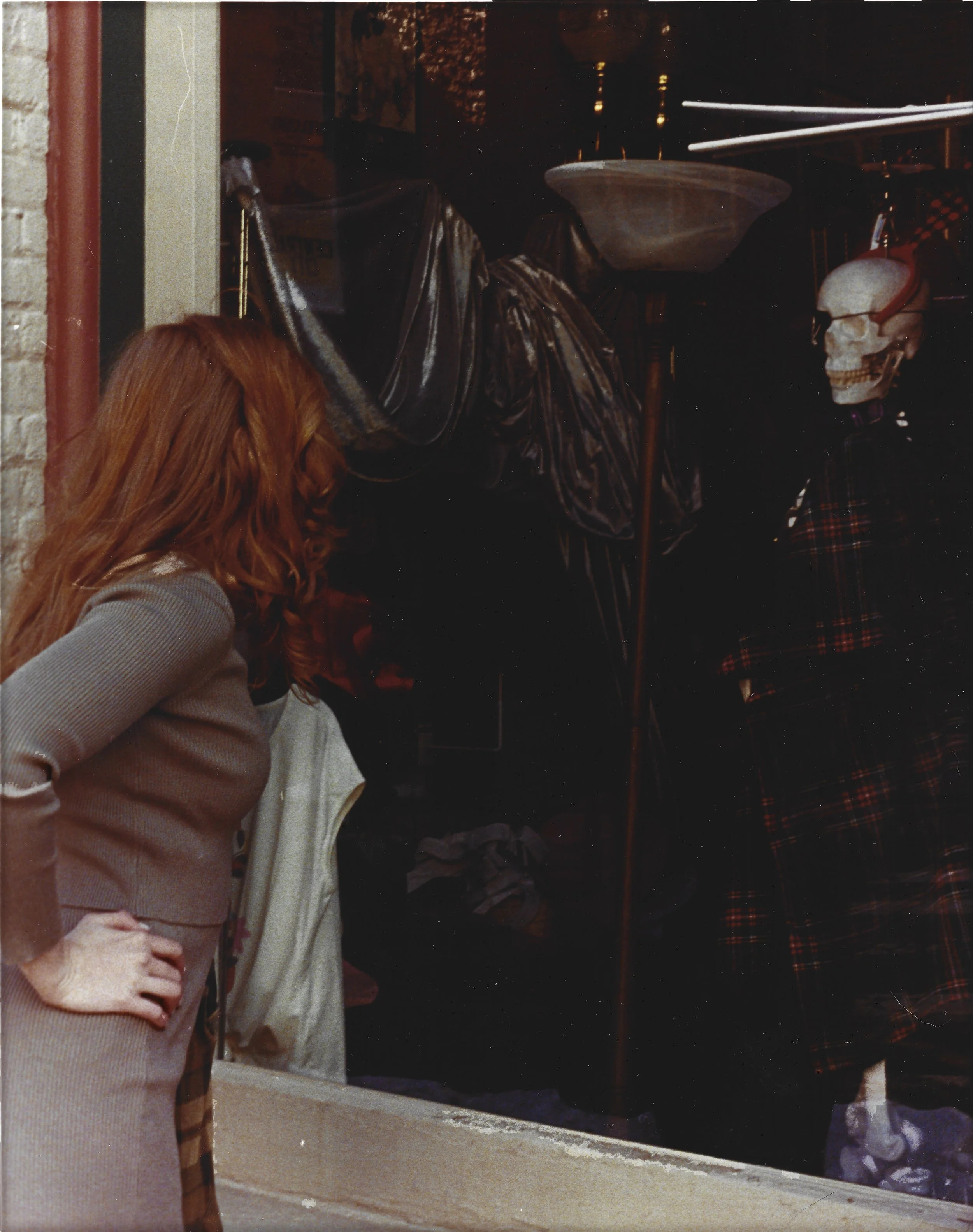 Woman with red hair looking at a store window display that includes a skeleton dressed in a plaid kilt and a headlamp, with a metallic bowl and dark fabric items inside the store.