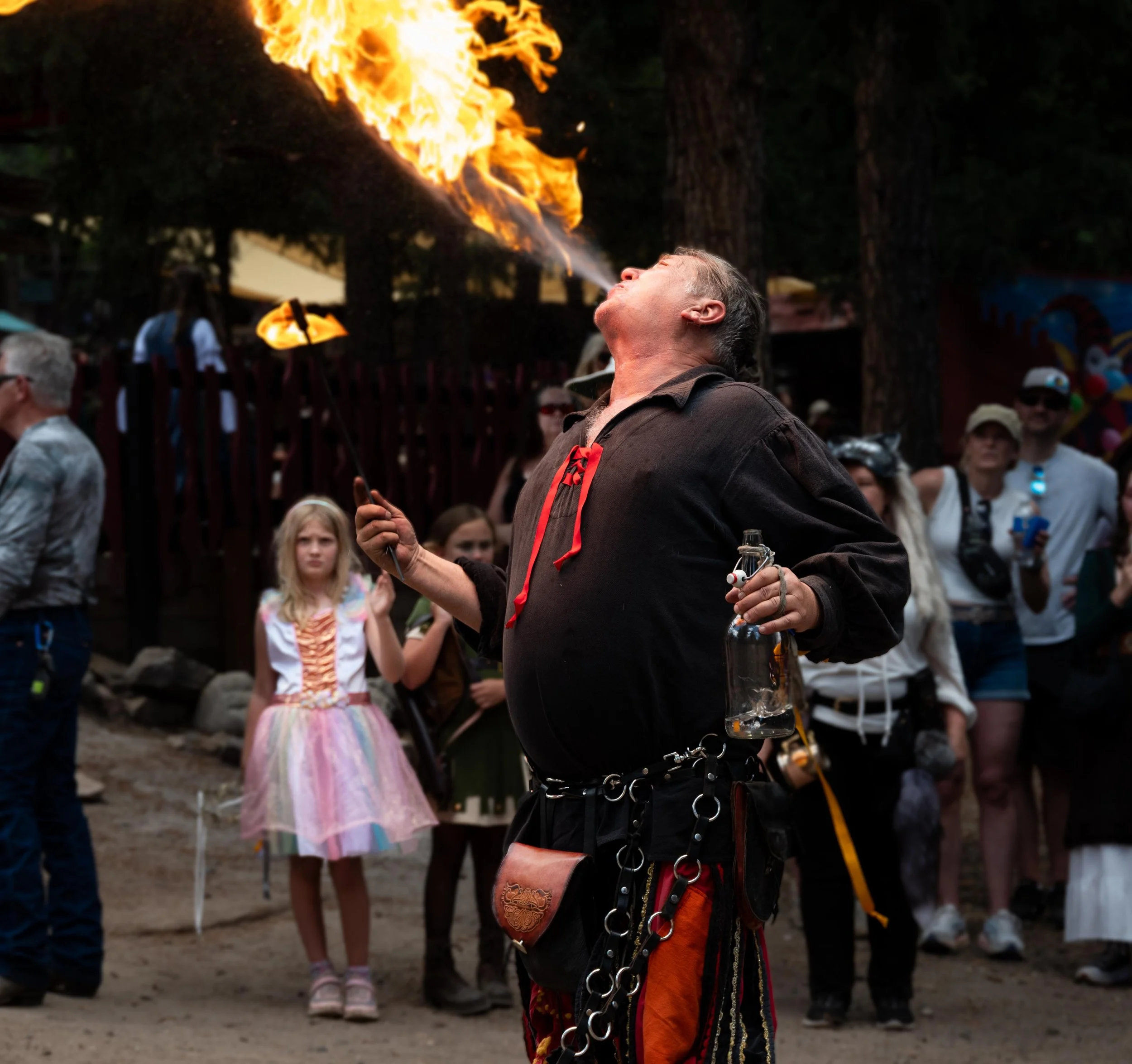 A fire performer in medieval-style clothing breathes fire at a crowded outdoor event, with onlookers watching.