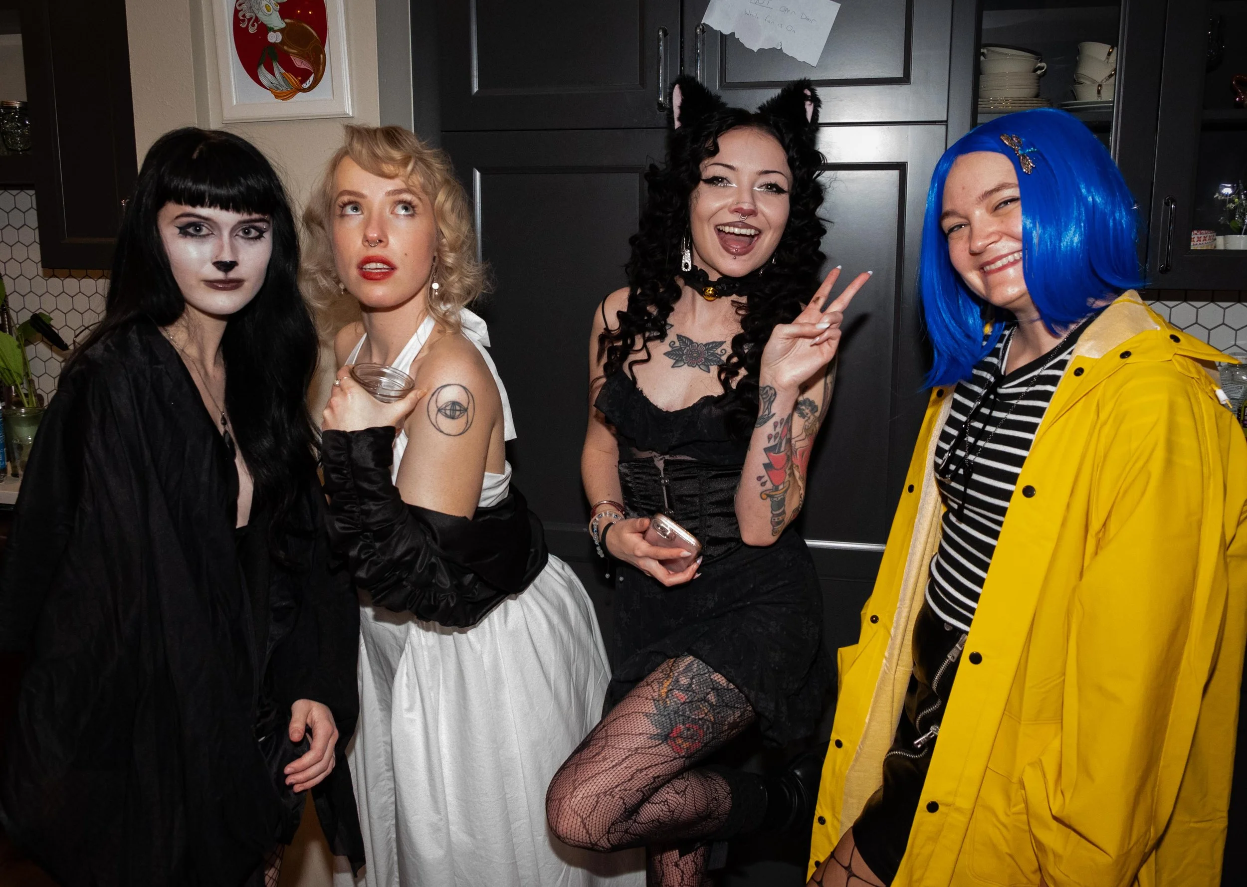 Four women in costumes pose together in a kitchen, dressed in dark and colorful outfits including a cat costume with fur ears, a vintage style dress, and a yellow raincoat, with one making a peace sign.