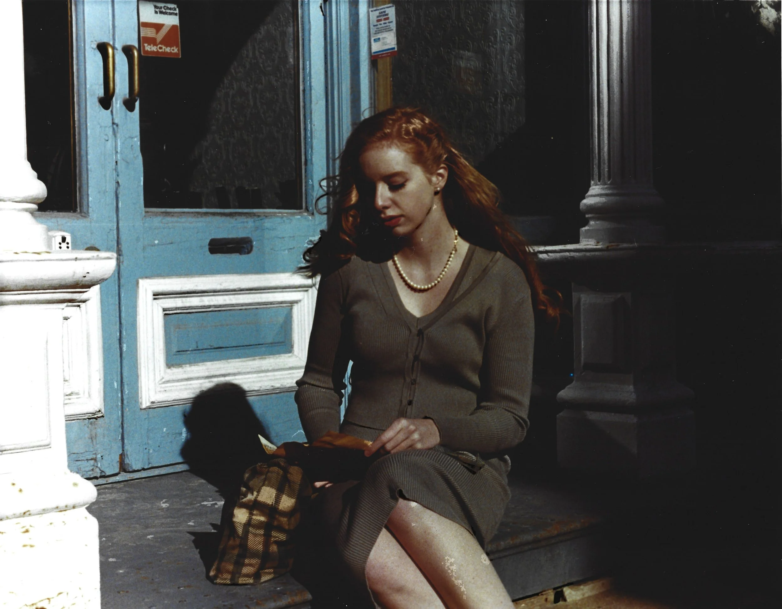 A young woman with red hair and fair skin sitting on a sidewalk at night, reading a book, with a blue door and a white column behind her.