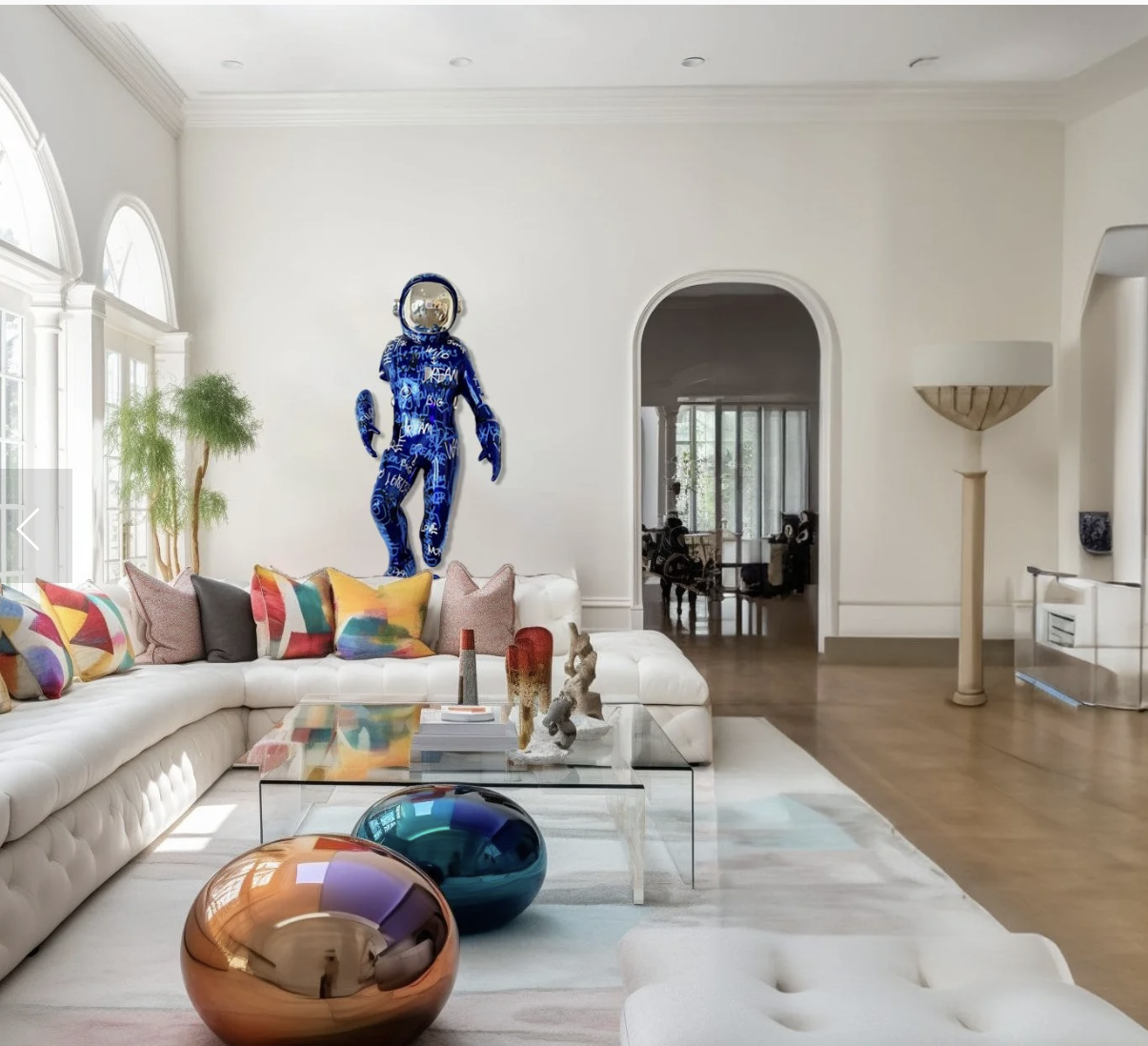 Living room with white sectional sofa, colorful pillows, glass coffee table, two large metallic decorative spheres, wall art of a humanoid figure with a mirror face dressed in a blue patterned suit, and a tall floor lamp.