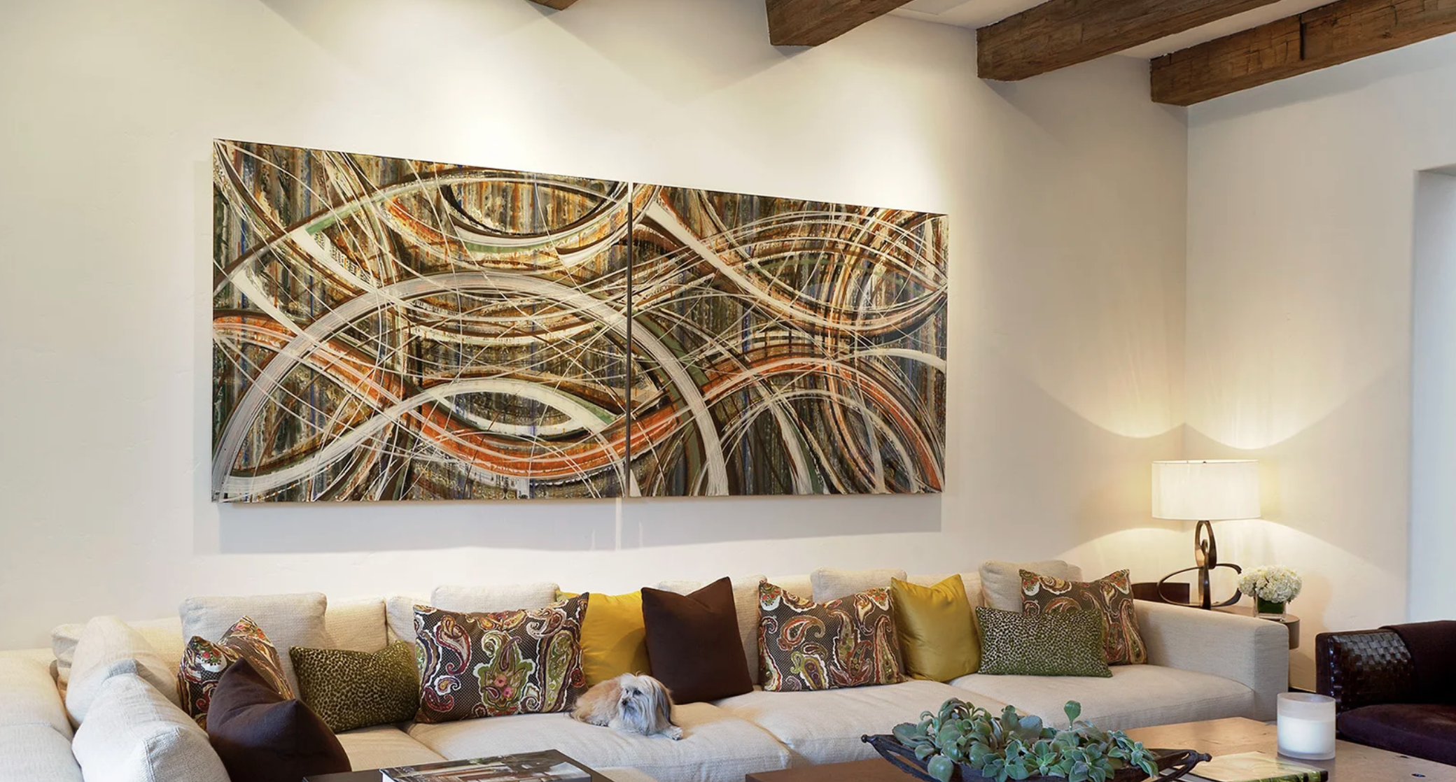 Living room with a large colorful abstract painting on the wall, beige sectional sofa with multiple decorative pillows, a small dog lying on the sofa, a table with a succulent plant, and a side table with a lamp and flower arrangement.