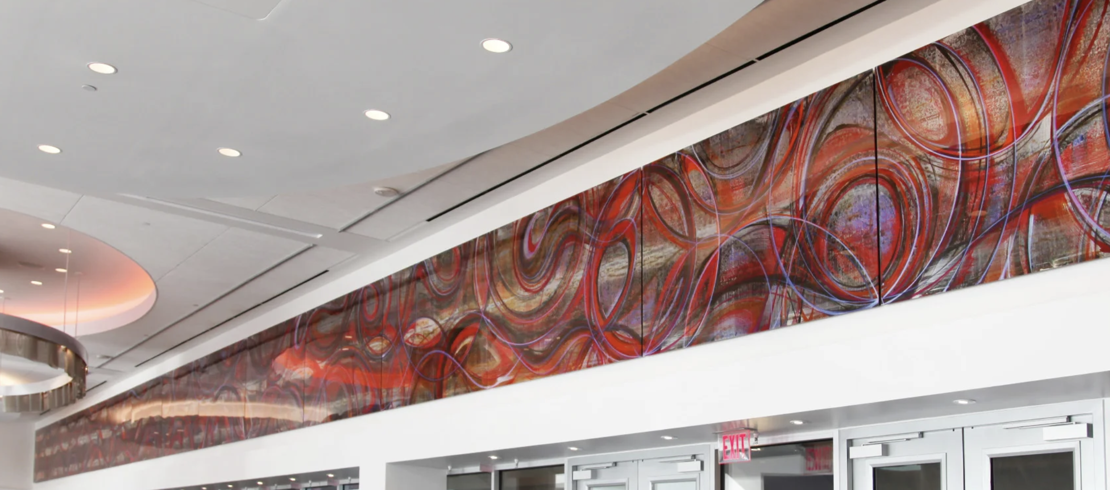 Interior view of a modern building lobby or hallway with a large, colorful abstract art installation on the wall, white ceiling with recessed lighting, and glass doors.