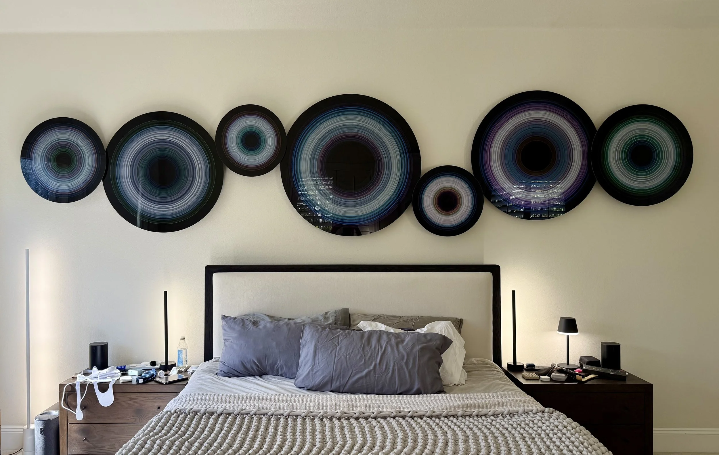 A bedroom with a bed, nightstands on both sides, decorative circular art hanging above, and various items on the nightstands including a water bottle, electronic devices, and lamps.