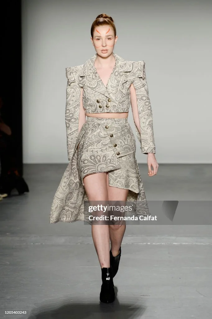 A model walking down a runway wearing a matching two-piece outfit with a paisley pattern, consisting of a cropped blazer with shoulder pads and a high-waisted skirt with button details, paired with black ankle boots.