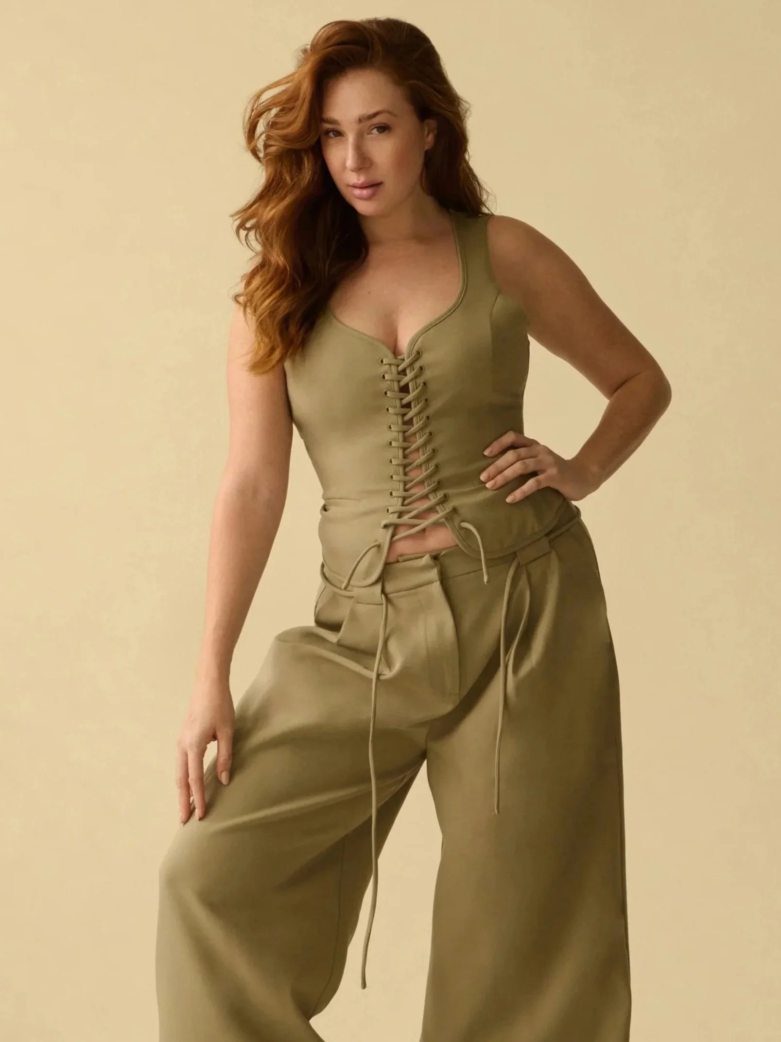 Woman with long red hair wearing a beige tank top with a lace-up front and beige wide-leg pants standing against a plain beige background.