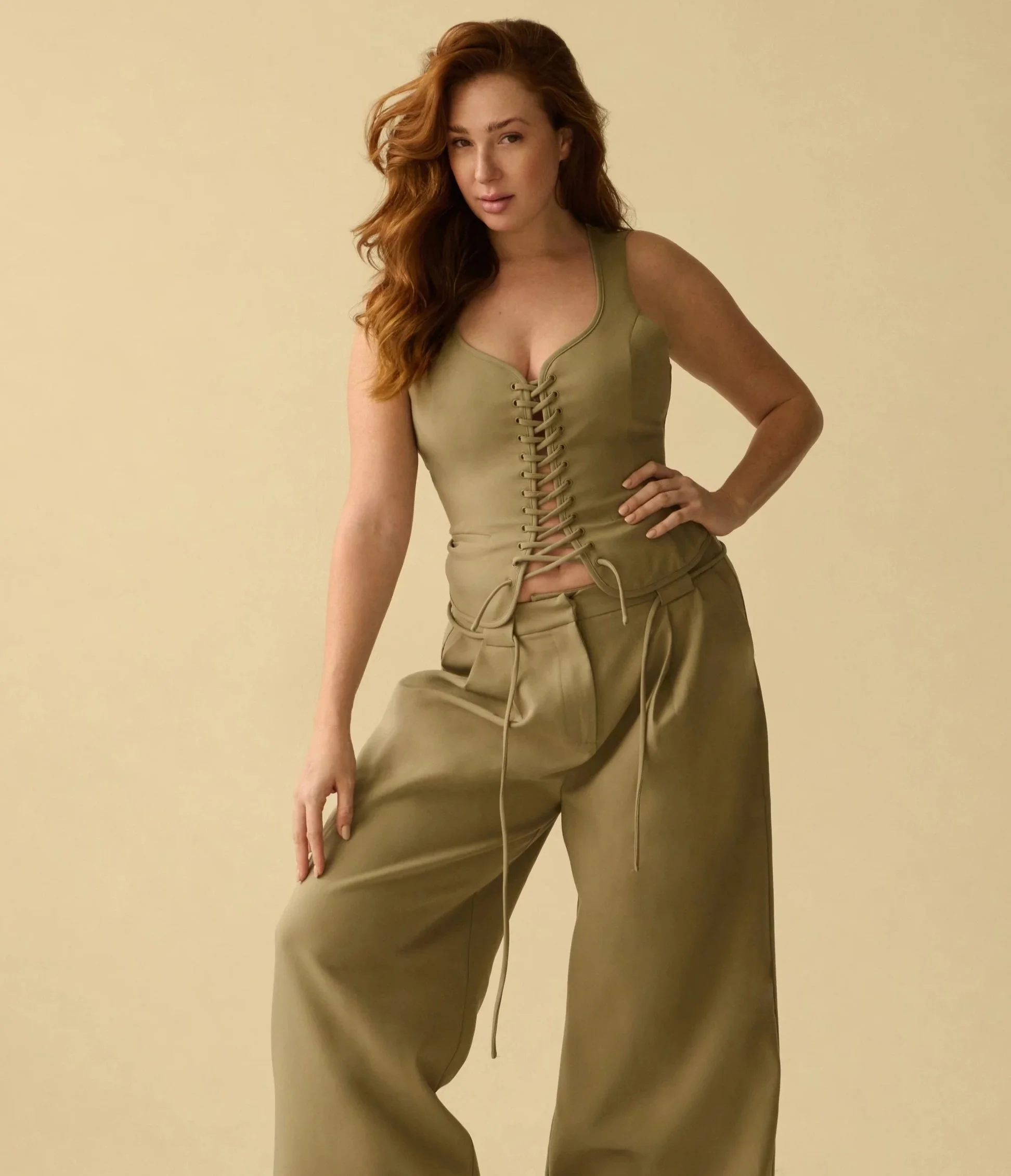 A woman with long red hair wearing a green tank top with lace-up front and matching wide-leg pants, posed against a plain beige background.